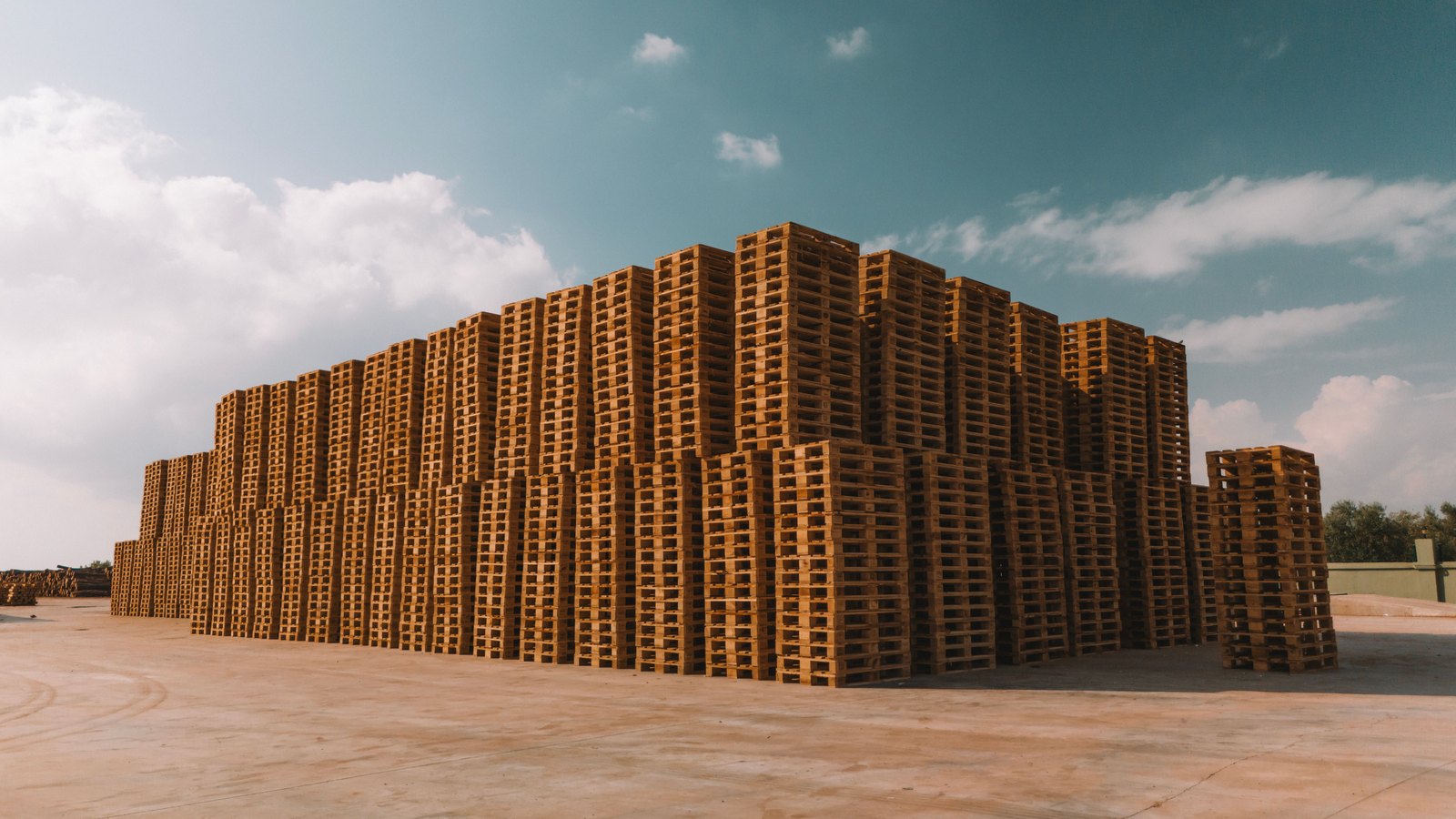 A very large block of bare wooden shipping pallets stacked several metres high in an open concrete yard, under a blue sky with scattered white cloud. No lettering, brand mark, vehicle or person is in the frame.