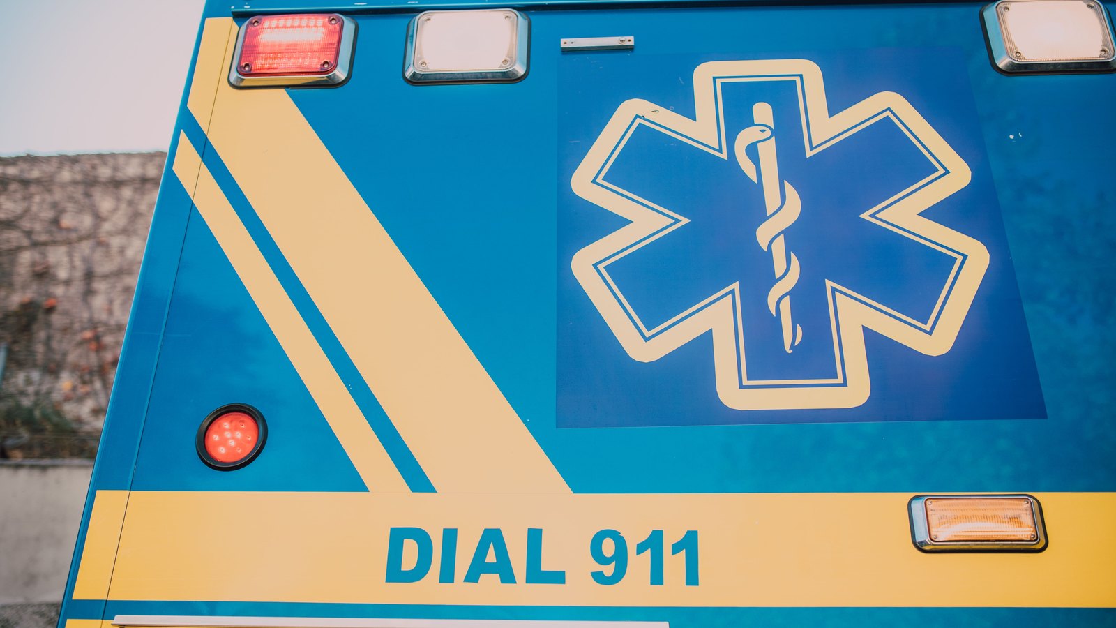 The rear panel of a blue and yellow ambulance carrying a star of life symbol and the words DIAL 911 in block capitals, with amber and red marker lights above and below it. No person, no face, no agency or department name, no address, no licence plate and no identifiable place appears in the frame. A stock photograph, not a photograph of this event.
