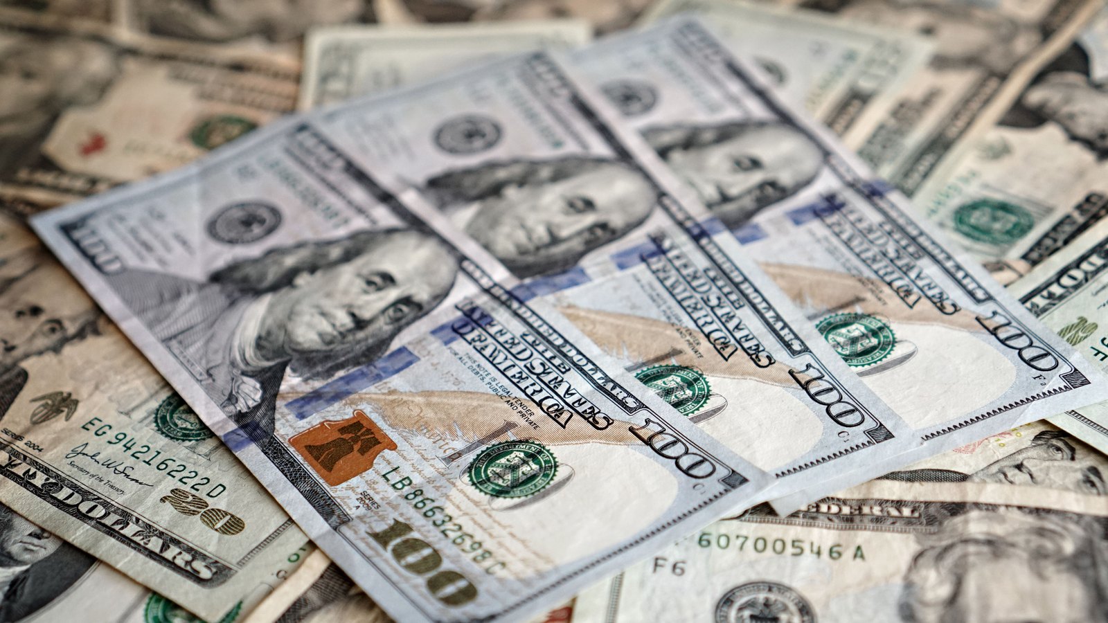 A loose pile of United States banknotes photographed close up, with three one hundred dollar bills lying face up across the top of the heap and older twenty and one hundred dollar notes underneath. Serial numbers and Treasury seals are visible. An illustrative photograph, not connected to this offering.