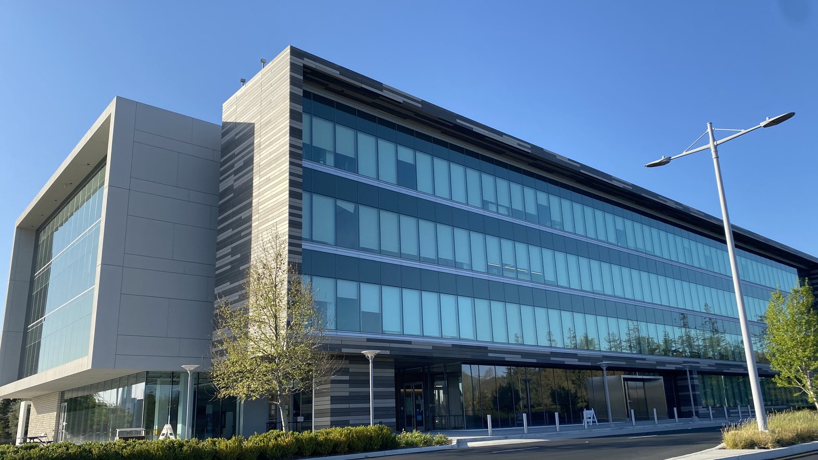 The Western Digital headquarters building in the Santa Teresa district of San Jose, California, a four storey office block with banded grey cladding and a full height glass curtain wall, seen from the street corner under a clear blue sky.