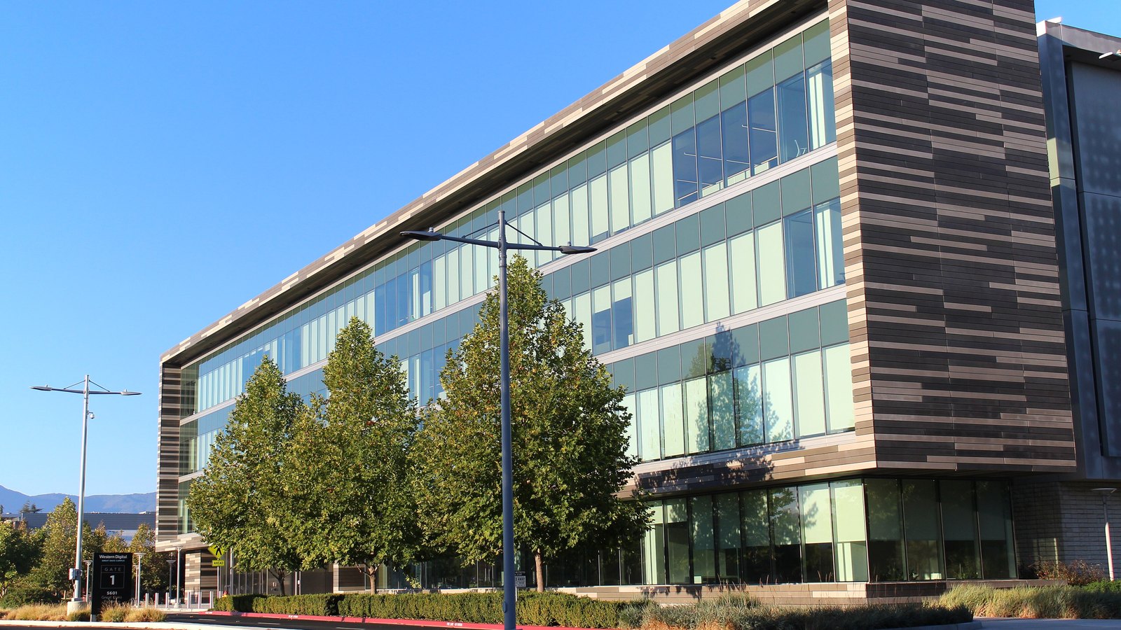 A four-storey glass and striped-panel office building at Western Digital's headquarters in San Jose, California, with a row of young trees and a street lamp in front under a clear blue sky.