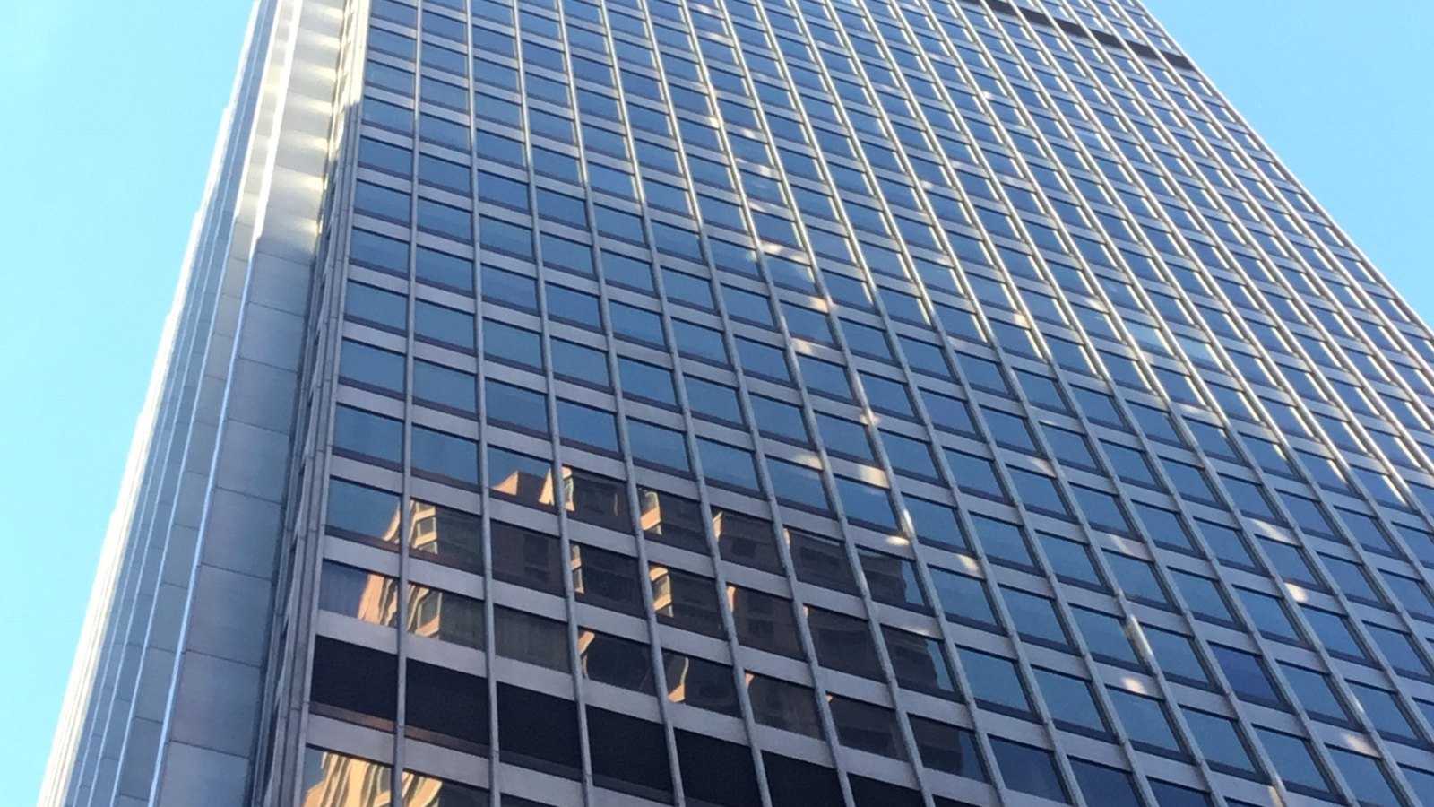 The glass and steel facade of 28 Liberty Street in Lower Manhattan, seen looking up from the pavement, with neighbouring buildings reflected in the lower windows.