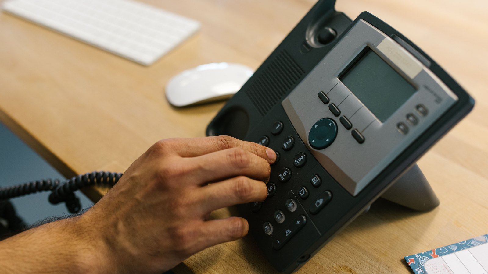 A close crop of a hand pressing the keypad of a black and grey desk telephone standing on a light wooden desk, with a computer keyboard and a white mouse out of focus behind it. No face, no place, no screen text and no logo is legible anywhere in the frame. It is a stock photograph and not a photograph of this event.