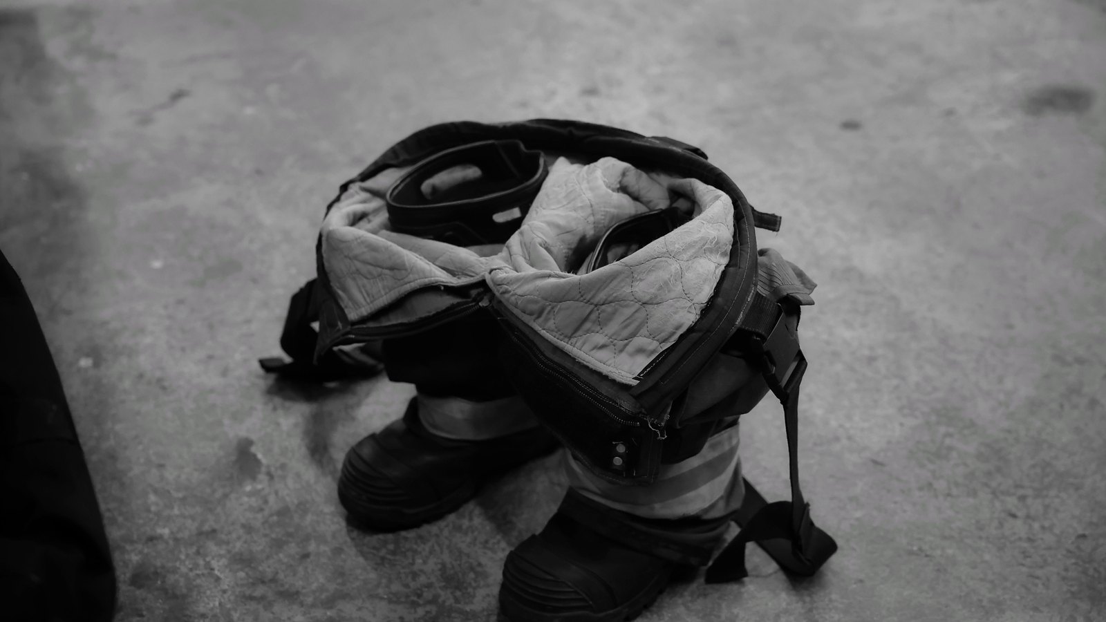 A black and white photograph of a firefighter's bunker trousers stood upright inside a pair of boots on a bare concrete floor, jacket collar folded open and shoulder straps hanging loose, ready to be stepped into. Apart from one person's trouser leg at the very edge of the frame there is nobody in the picture, and there is no text, no badge, no vehicle, no number plate, no building and no department or agency marking anywhere in it. Stock photograph, not a photograph of this event.
