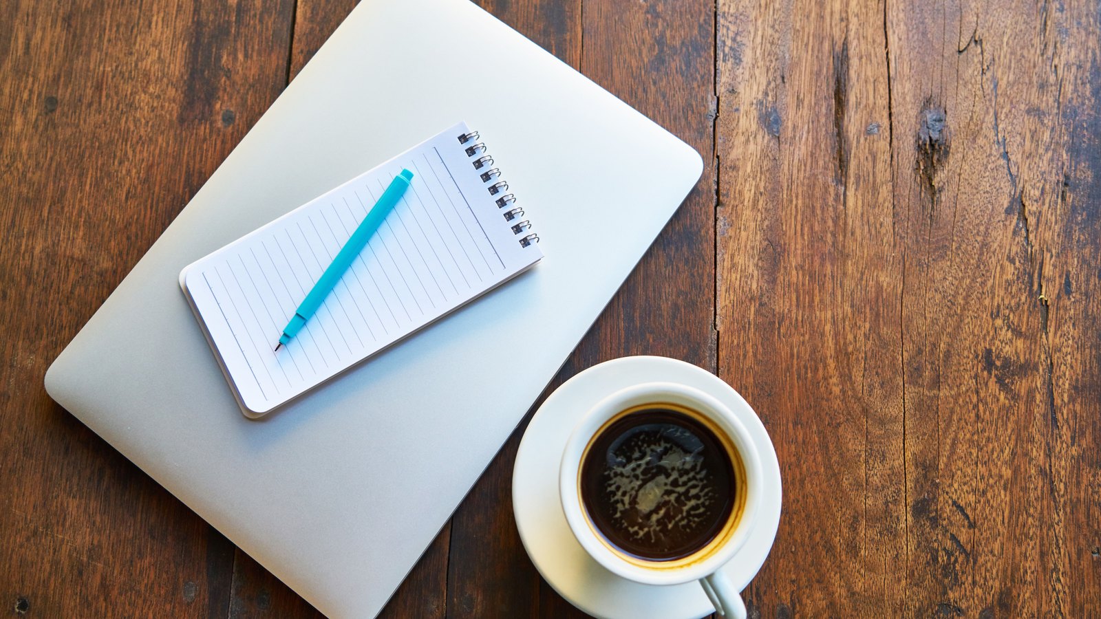 A blank notepad, a pen, a closed laptop and a cup of coffee on a wooden table, used as a neutral illustration. It shows nobody and nothing connected to this case.