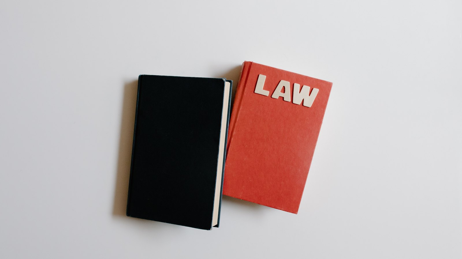 Two closed hardback books lying flat on a plain pale grey surface, one black and one red, the red one spelling the word LAW in raised wooden letters, with no people or setting in view.