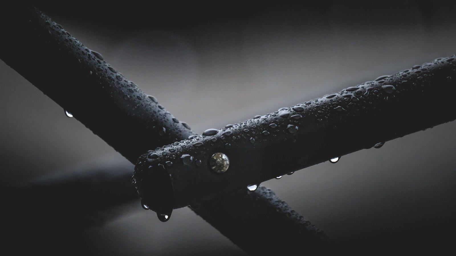 Two dark metal bars crossing at a bolted joint, beaded with rainwater, against an unlit grey background.