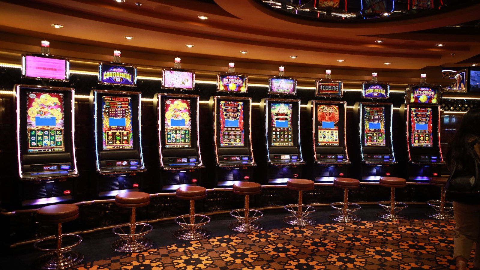 A row of eight lit slot machines against a dark wall, with empty leather topped stools in front of them and a patterned carpet in the foreground. A generic illustration, not the Collier County locations.