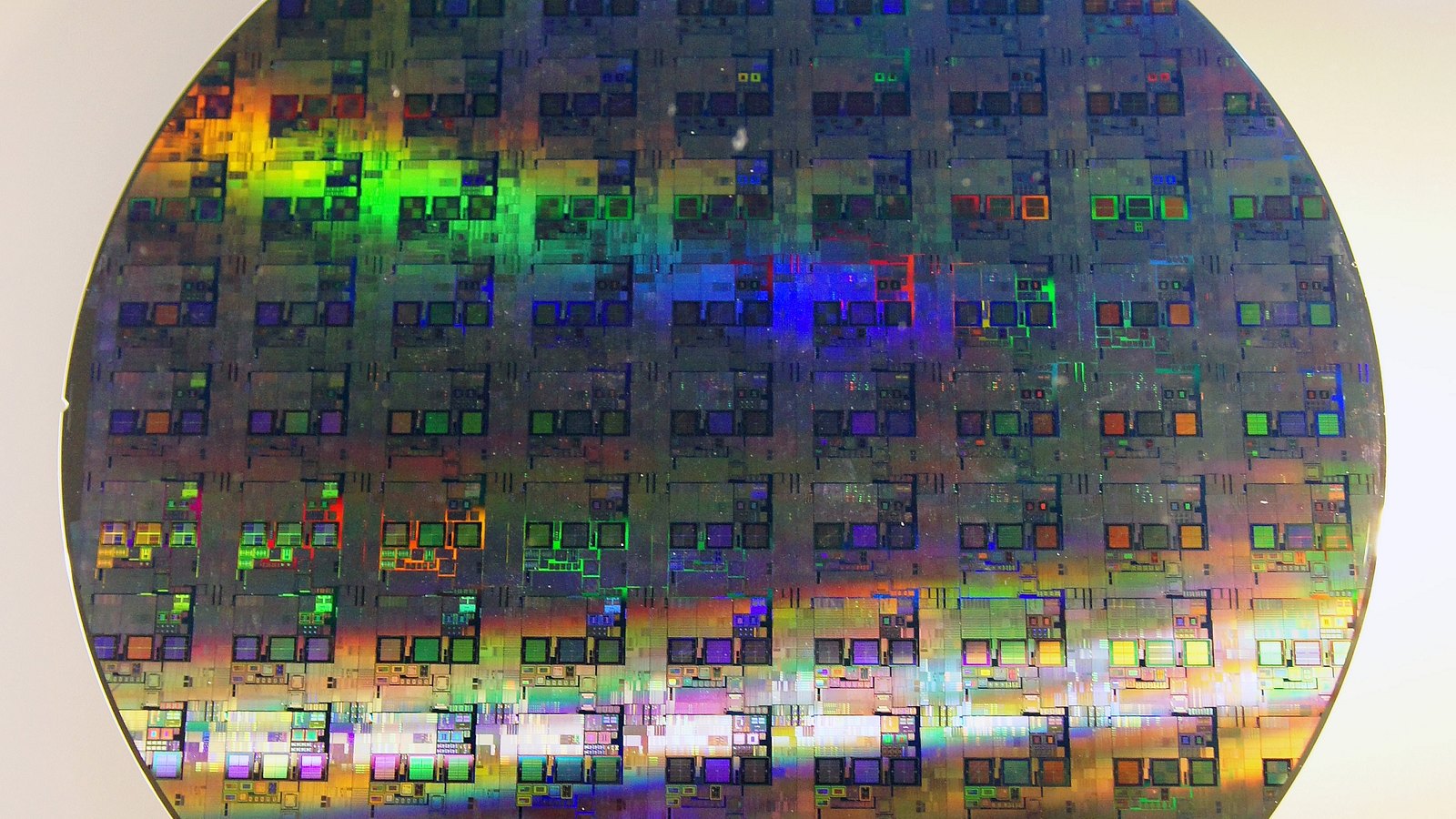 A 12-inch silicon wafer photographed flat against a pale background, its surface covered in a regular grid of rectangular integrated circuit dies and shot through with bands of rainbow interference colour. An illustrative photograph of a semiconductor wafer, not one of this company's own.