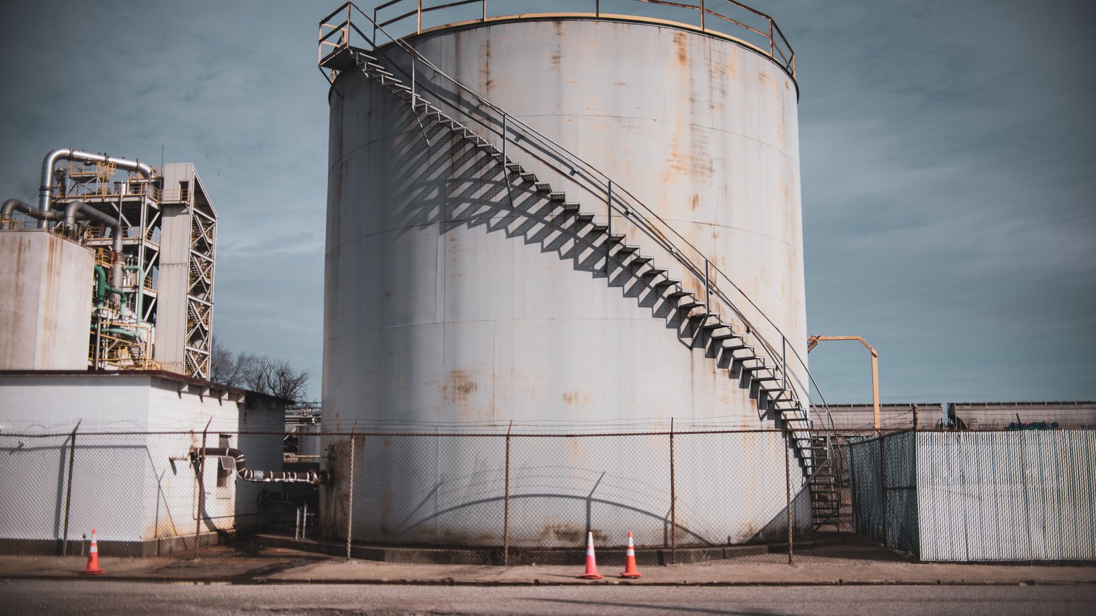 A large pale bulk storage tank with an external stairway spiralling up its side, behind a chain-link fence topped with barbed wire, with a smaller process unit and insulated pipework to the left and three traffic cones on the road in front.