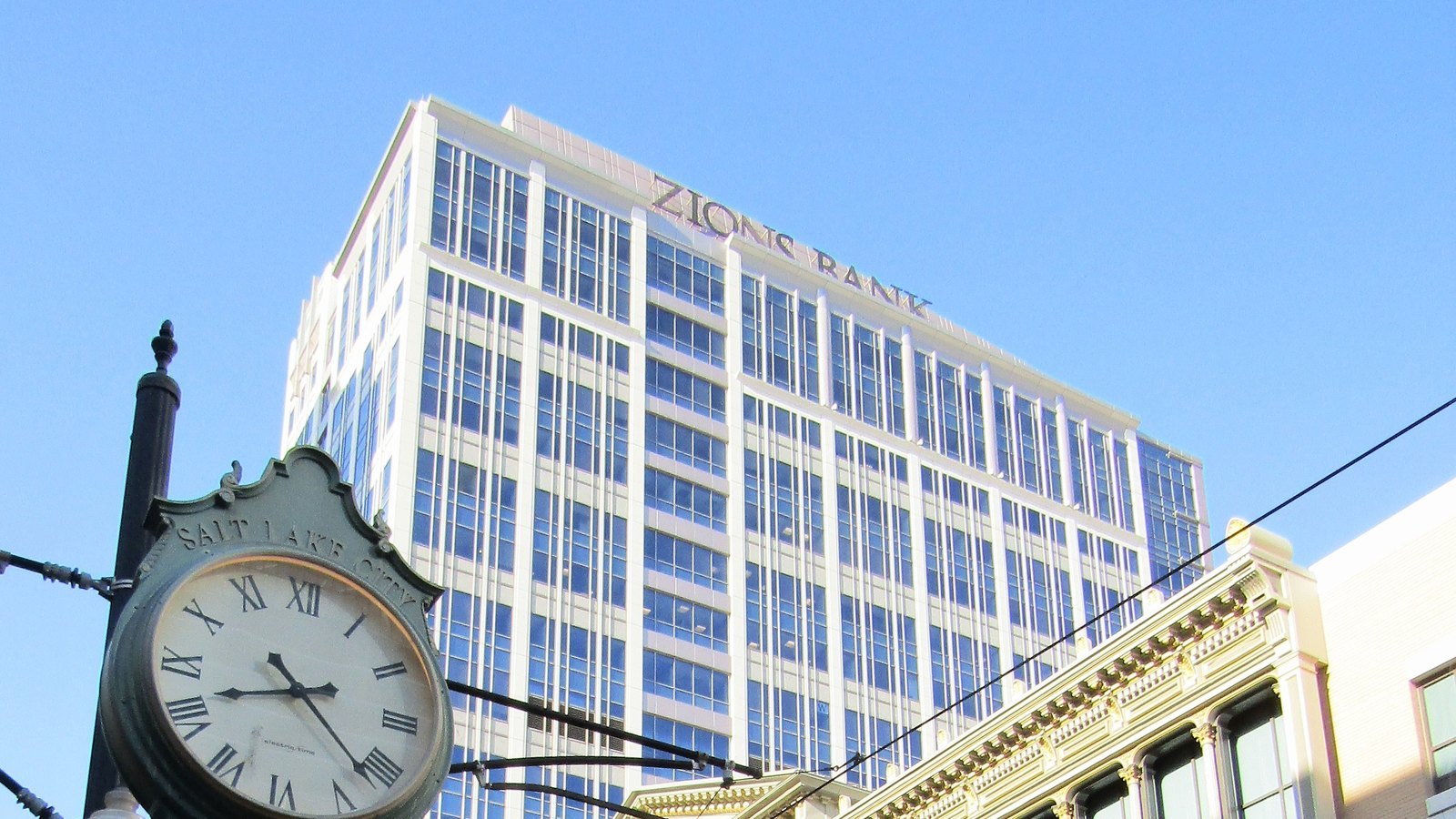 The upper floors of the Zions Bank tower in Salt Lake City, its name in lettering along the roofline, behind an ornate green street clock and the cornice of an older building.