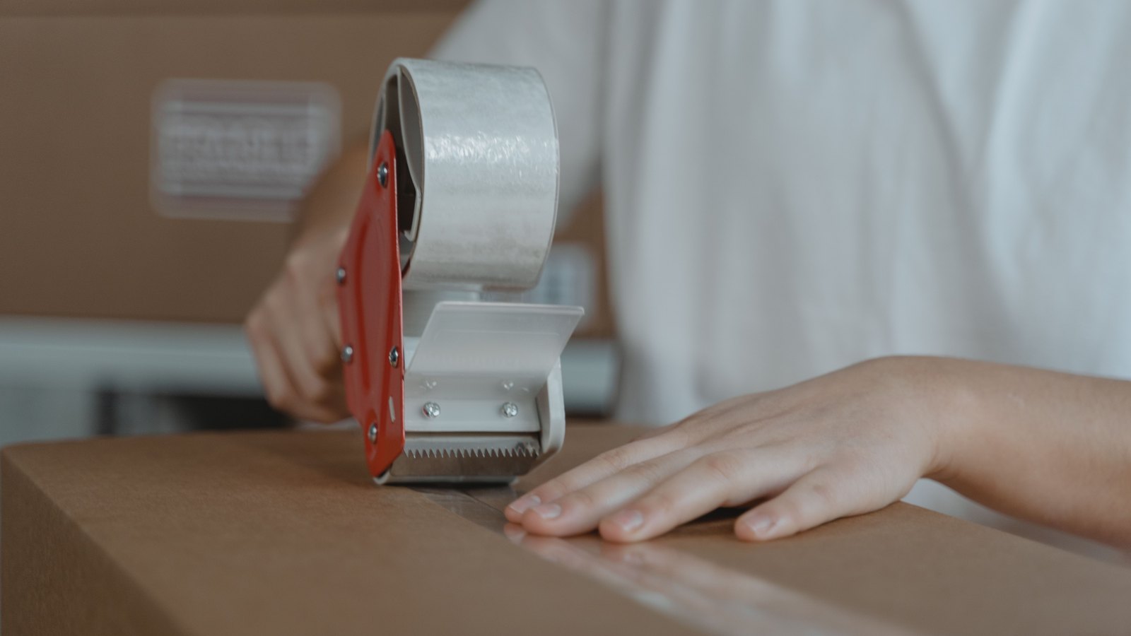 A person in a white t-shirt, shown from the shoulders down, running a red handheld tape gun along the seam of a sealed brown cardboard box, with more flattened boxes stacked behind.