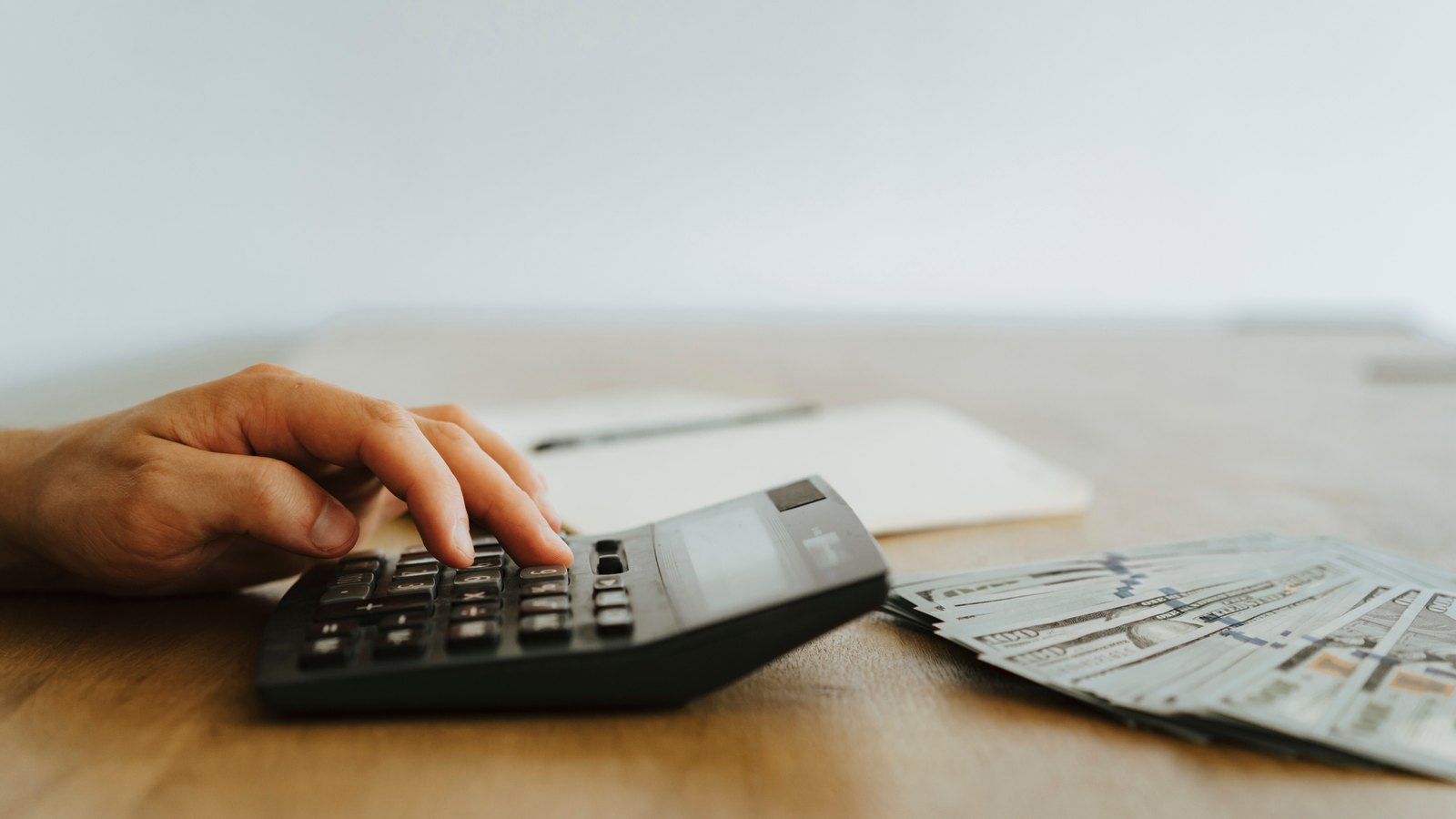 Hands using a calculator beside United States banknotes on a desk.