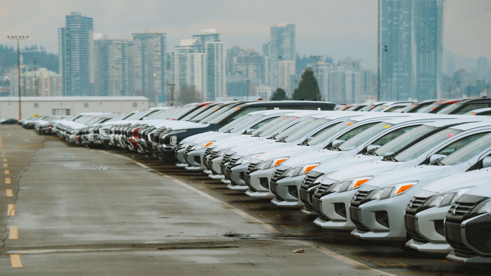 A long row of new vehicles parked nose-out on a wet lot under flat grey cloud, with distant high-rise towers behind.
