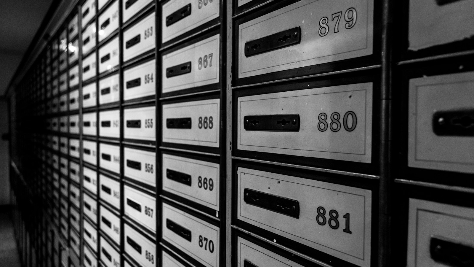 A long wall of small metal mailboxes in an apartment building, photographed in black and white at a sharp angle so the numbered doors recede into shadow.