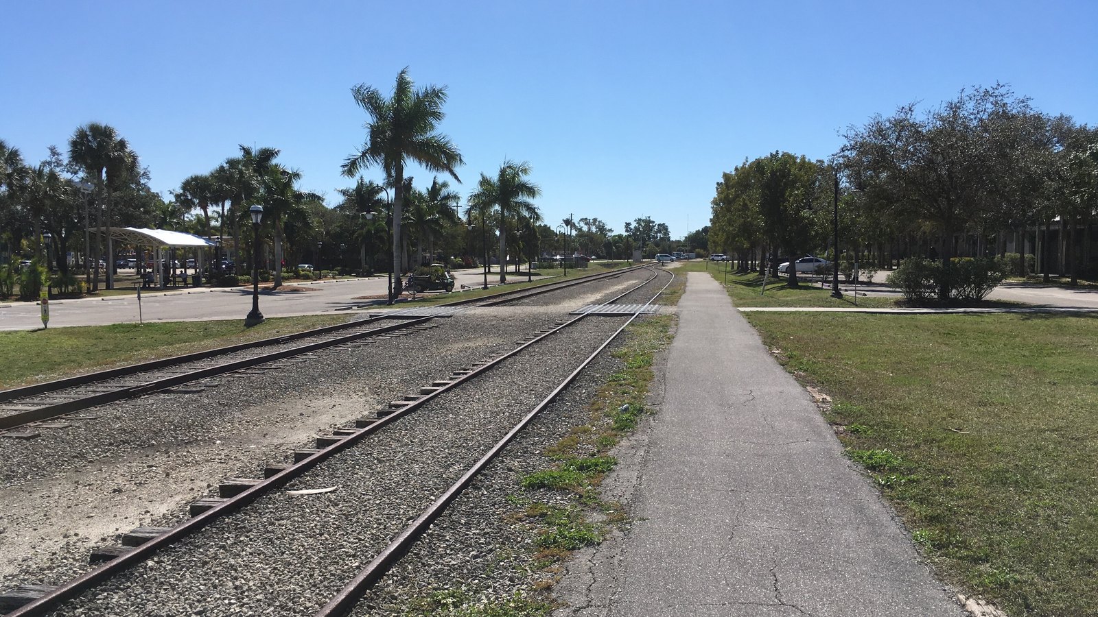 Railroad tracks curve through downtown Bonita Springs beside a palm-lined park and path.