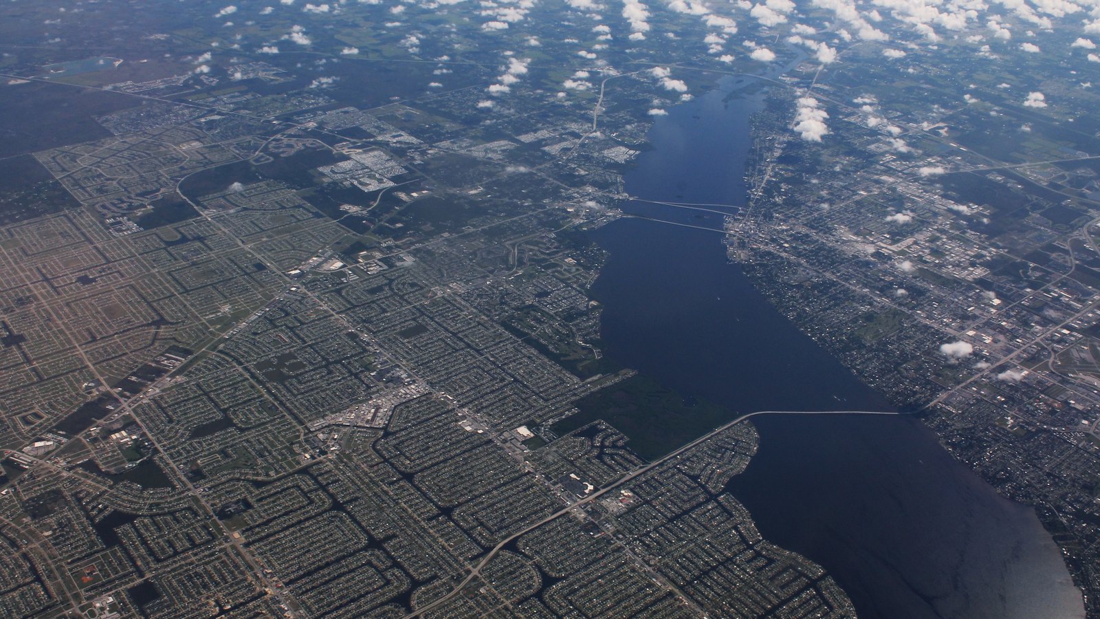 A high-altitude aerial photograph of a wide grid of low-rise residential streets and canals on one bank of a broad river, with bridges crossing to a denser built-up bank opposite, under scattered cumulus cloud.