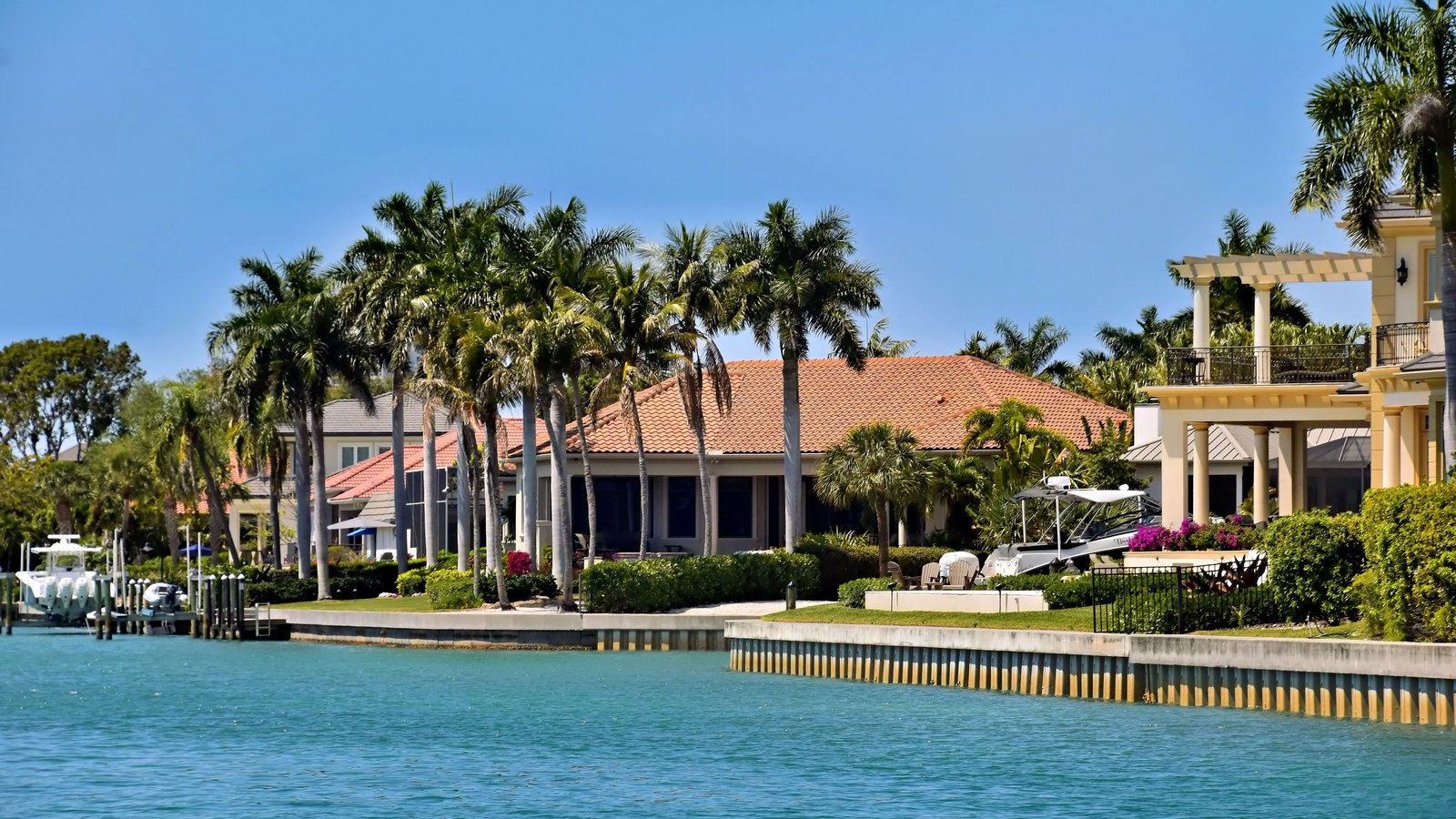 A row of single-story and two-story waterfront houses with barrel tile roofs standing behind royal palms, concrete seawalls and private boat docks on a calm turquoise canal under a clear sky.