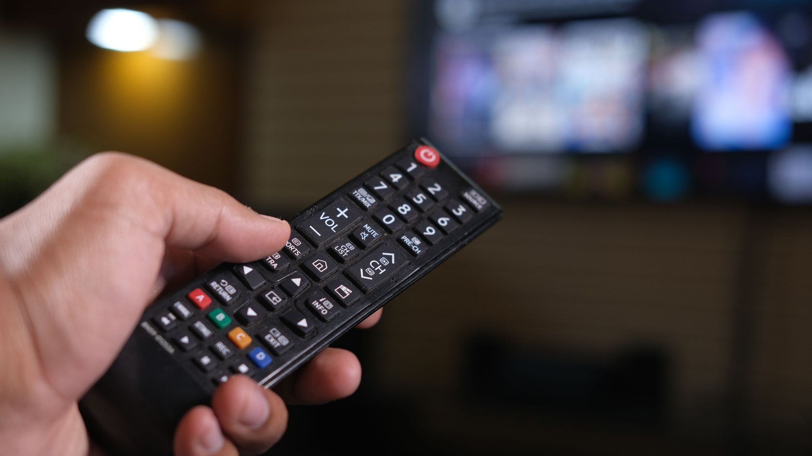 A hand holding a television remote control, with a blurred grid of on-screen content tiles behind it.