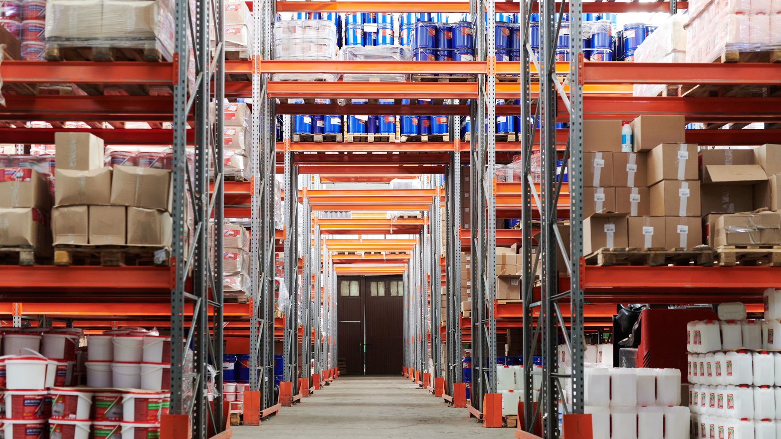Wide aisle in a warehouse with pallet racking stacked full of boxes, drums and containers on both sides