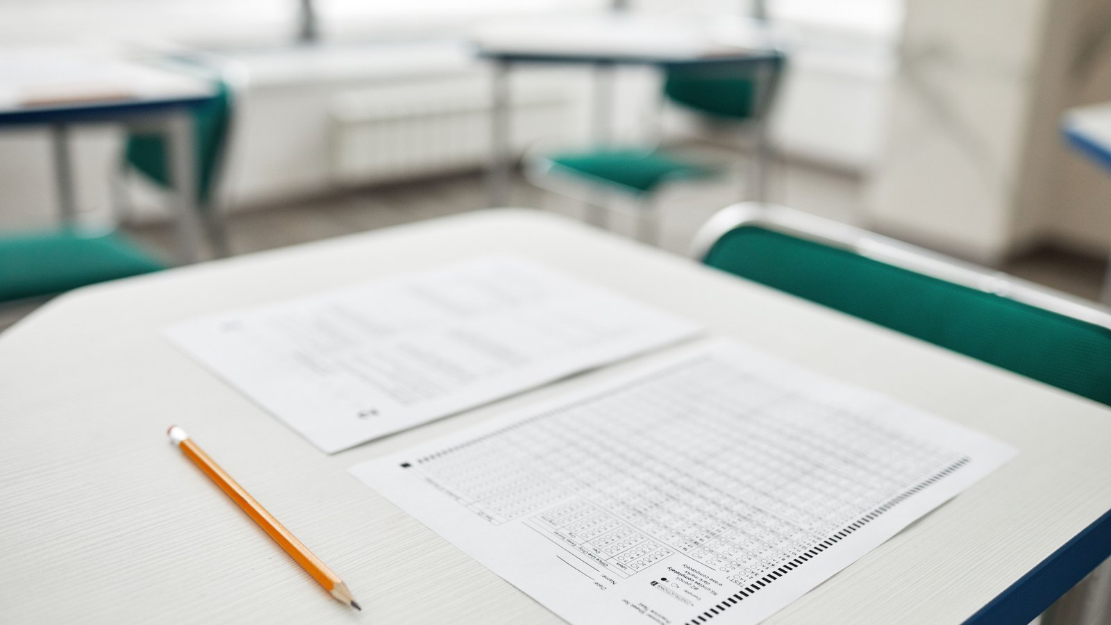 A multiple-choice answer sheet and a question paper lie beside a pencil on an empty classroom desk, with more desks out of focus behind.