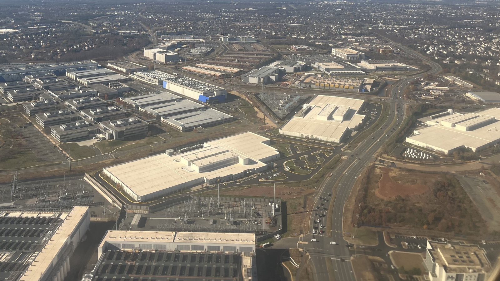 Aerial view of large white-roofed data center buildings, electrical substations and a divided highway in Loudoun County, Virginia, near Ashburn, with housing developments behind.