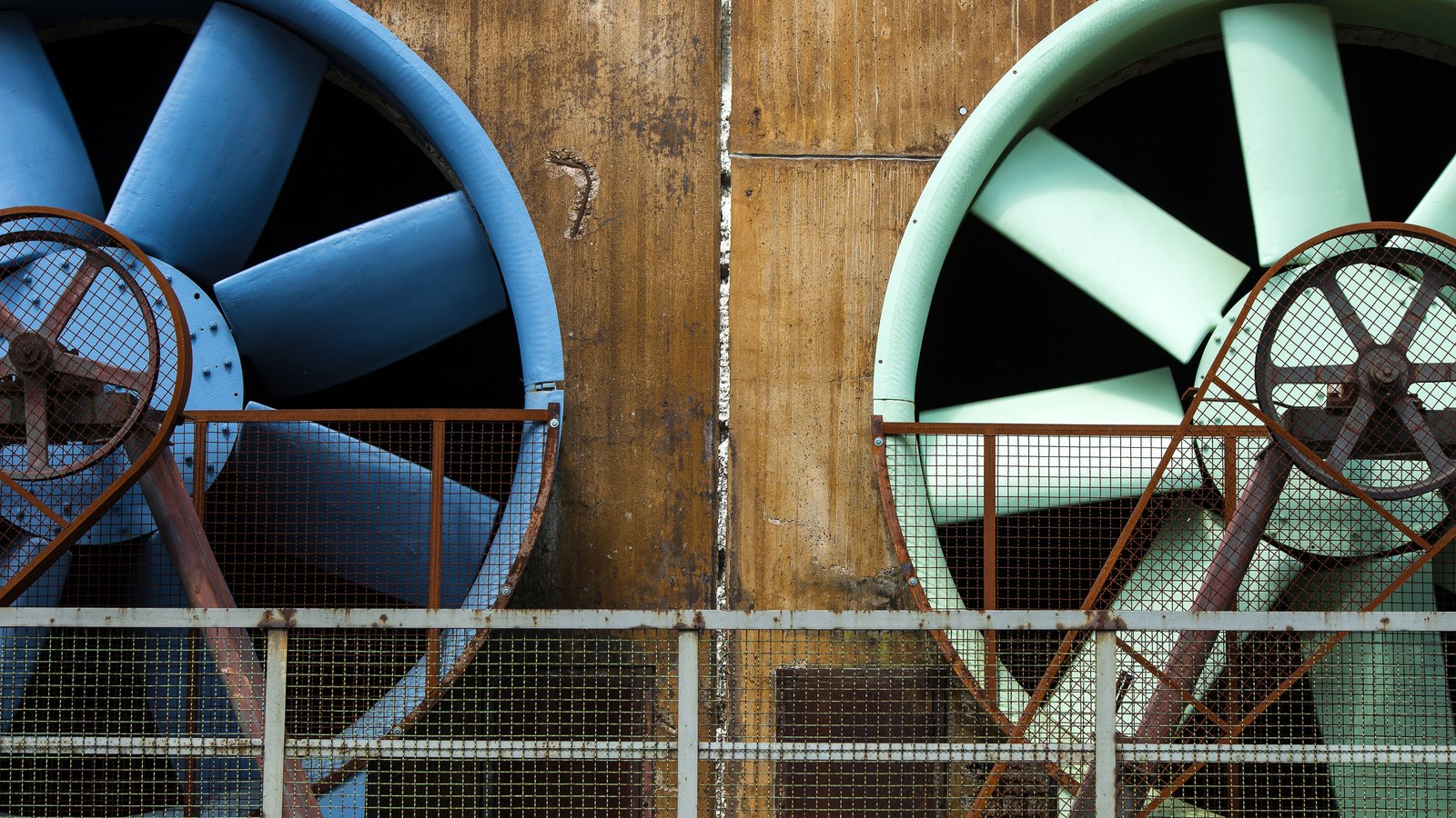 Two large industrial cooling tower fans set into a weathered concrete wall behind a metal railing, one blue and one pale green.