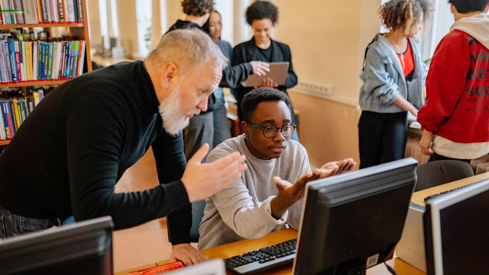 A teacher helps a student use a desktop computer in a library while other students work nearby.