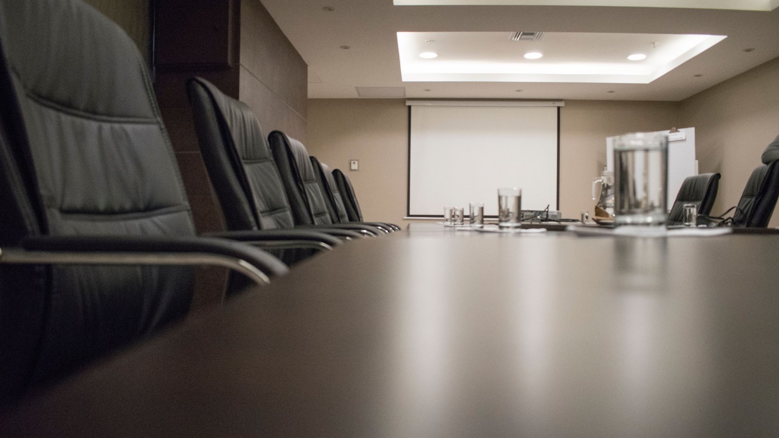 A low view along an empty boardroom table, high-backed black leather chairs down one side, a water pitcher and glasses at the far end in front of a blank projection screen.