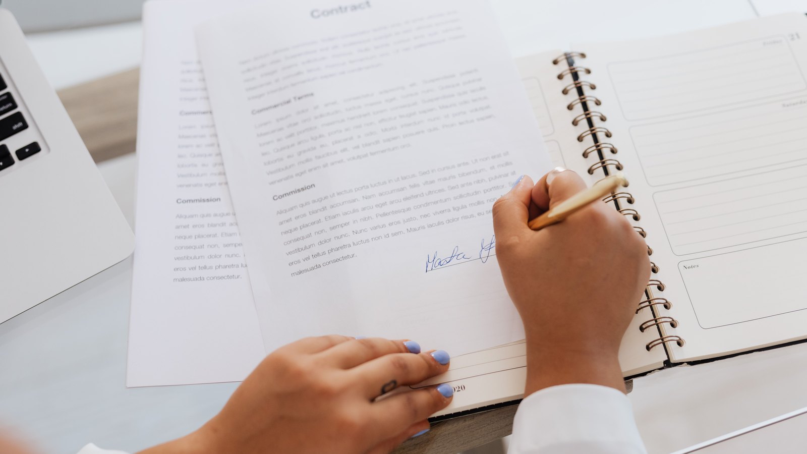 Two hands at a desk, one steadying a printed contract carrying a signature, the other writing in a spiral notebook beside a laptop.