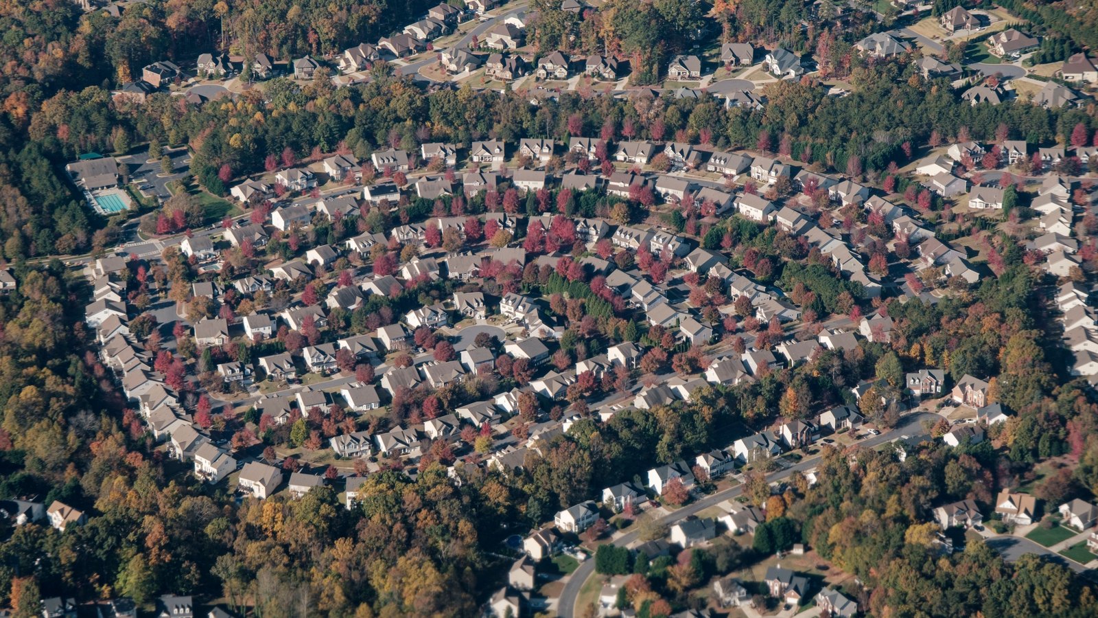 An aerial photograph of a suburban housing development, rows of similar pitched roofs following concentric curved streets between wooded lots, autumn colour in the trees.