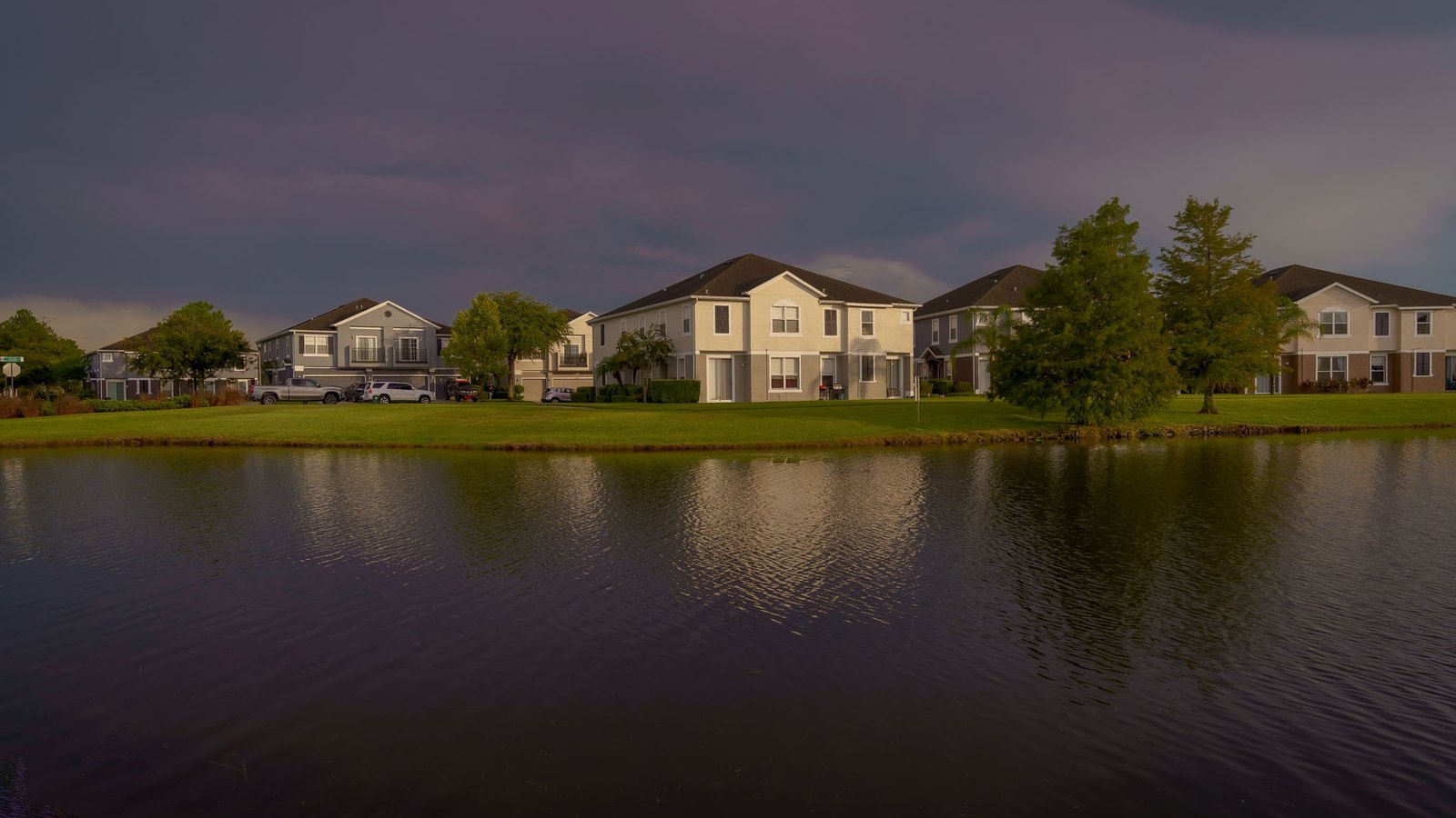 Two-story stucco houses behind a retention pond at dusk in a Florida suburb, with a bald cypress tree and warm light on the homes under a passing storm sky.