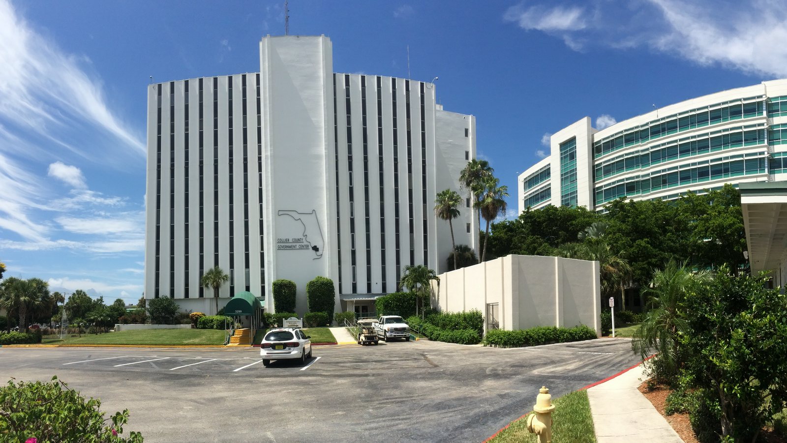 The white tower of the Collier County Government Center in Naples, with the Florida outline and county name on its face, beside the curved glass courthouse annex under a blue sky.