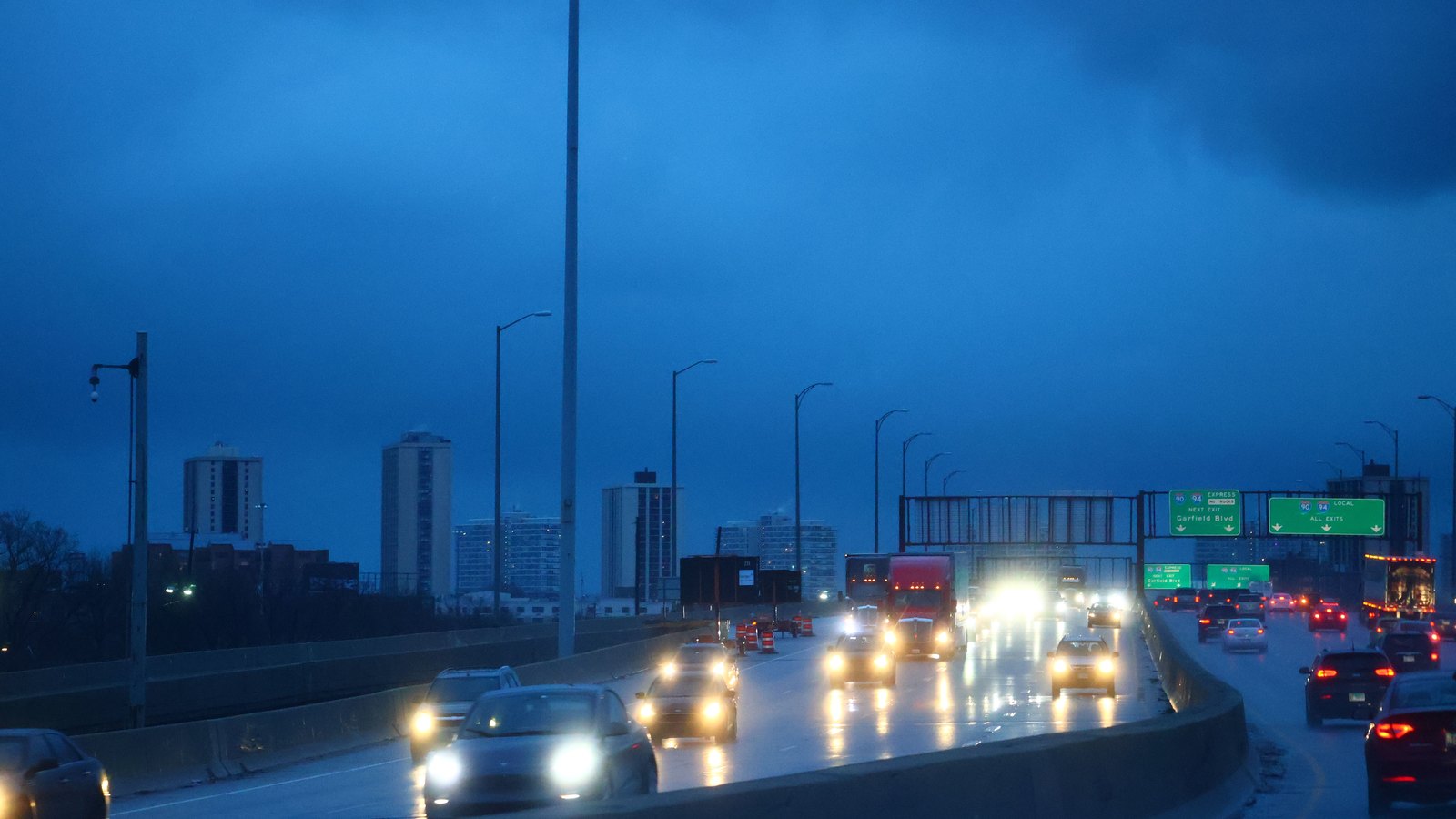 Headlights and taillights on a wet multi-lane interstate at dusk, a city skyline low on the horizon behind.