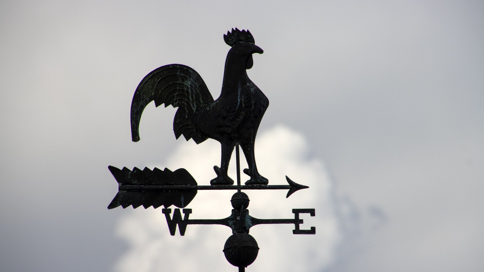 A rooster weather vane silhouetted against a clouded sky, its arrow pointing west.