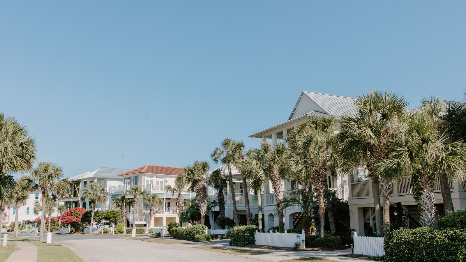 A quiet coastal residential street lined with sabal palms, low white picket fences and pale raised houses with metal roofs under a clear sky.
