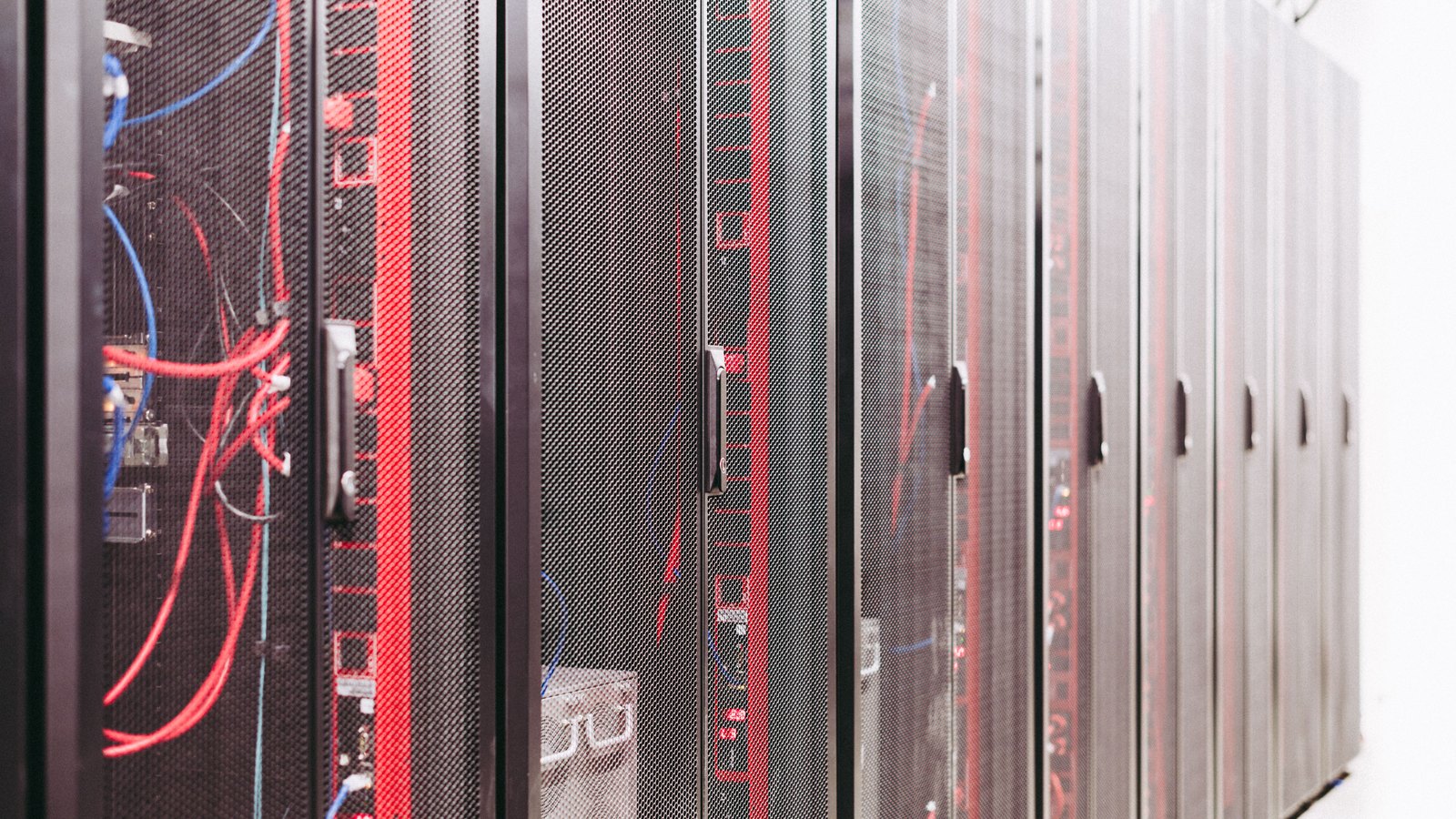 Rows of black server cabinets with perforated mesh doors in a data hall, red and blue patch cables visible inside the nearest racks.