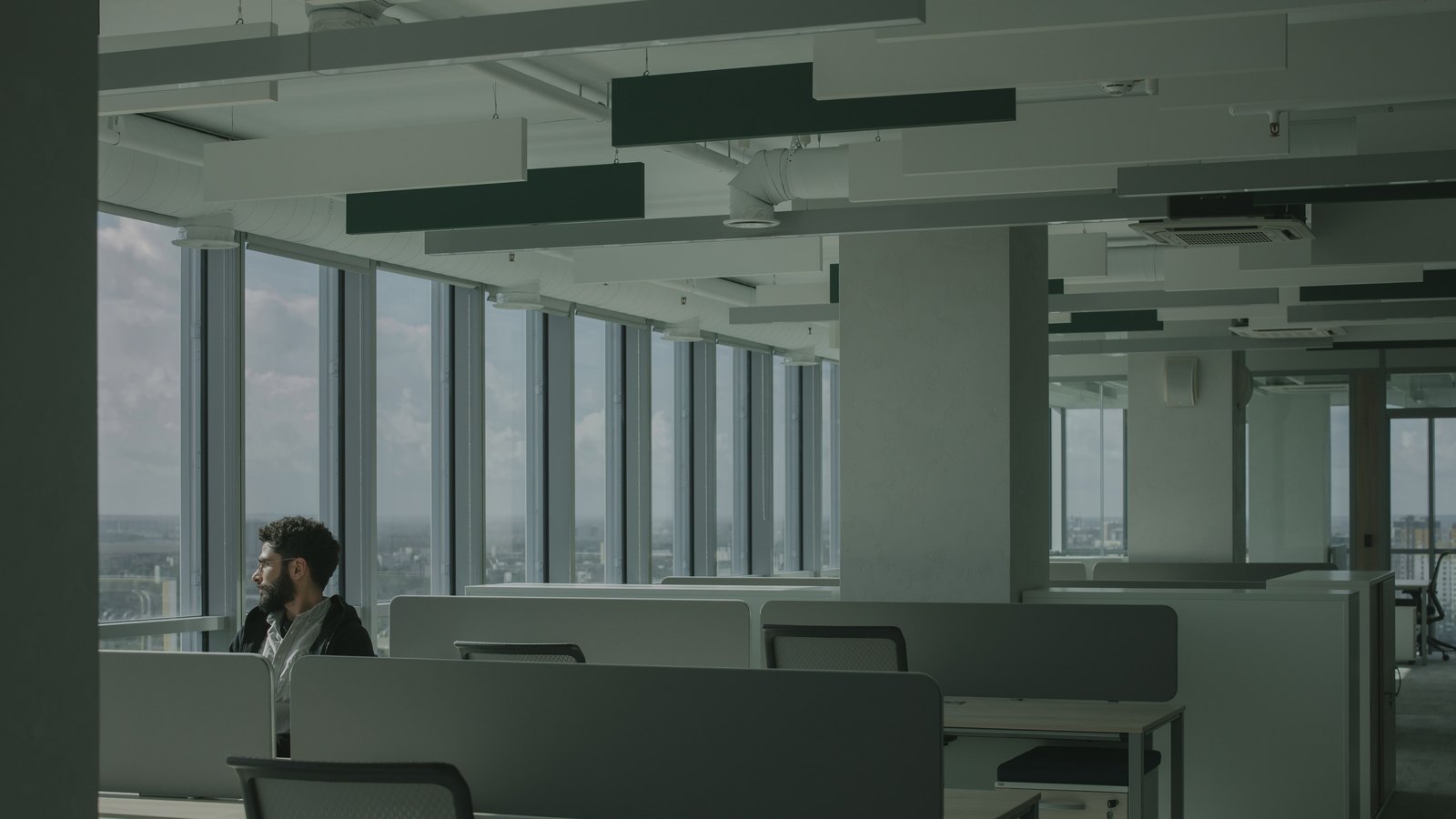 One person seated at a desk on a large open plan office floor, the surrounding rows of desks empty, under flat daylight from a window wall.