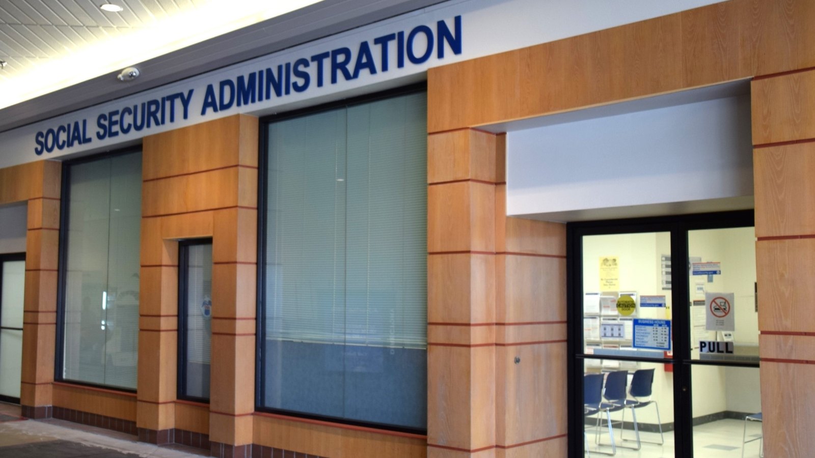 The entrance to a Social Security Administration field office, with the agency name in blue lettering above the windows and a waiting area visible through the glass door.