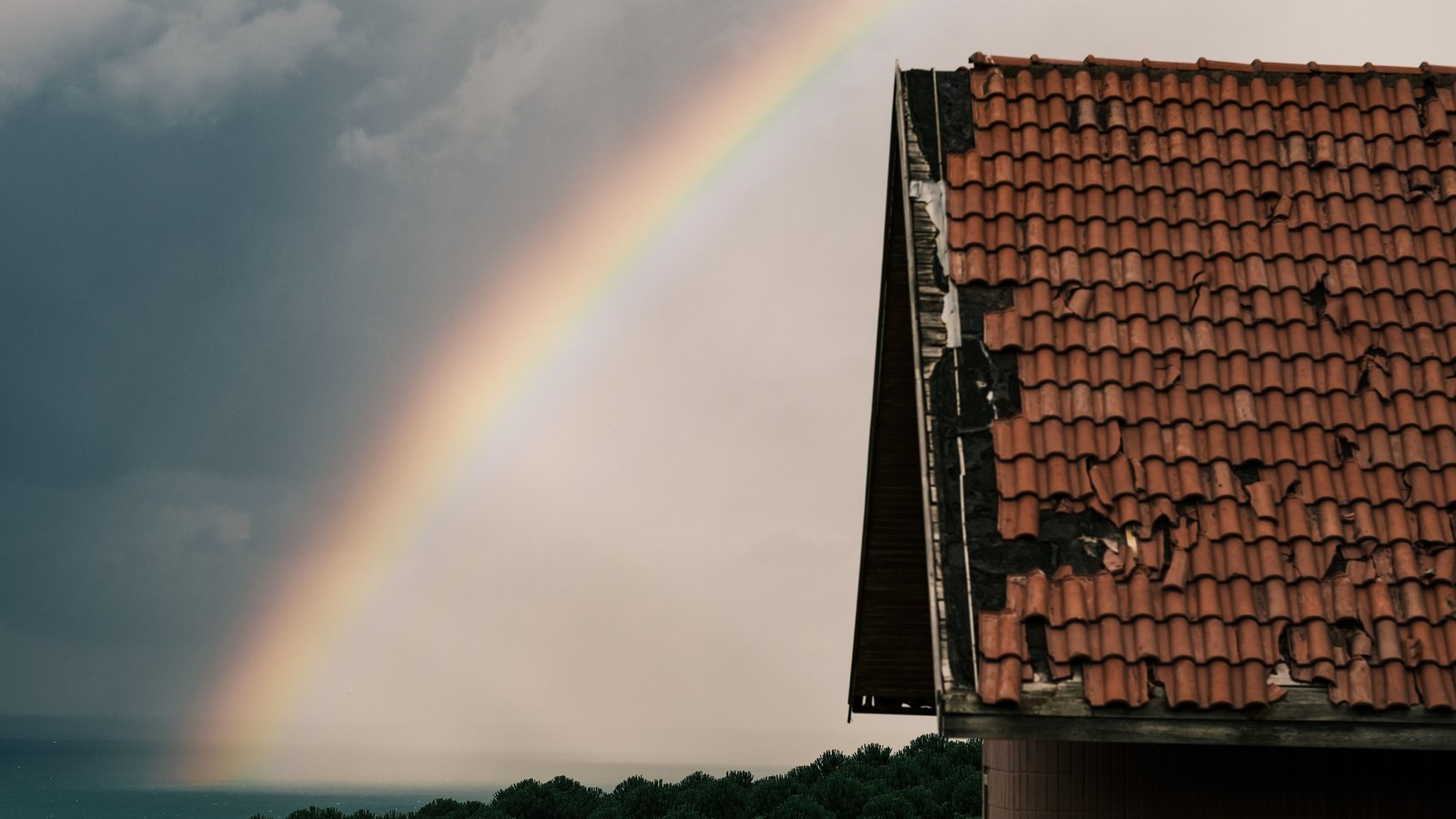 A damaged terracotta tile roof beside the sea beneath storm clouds and a rainbow