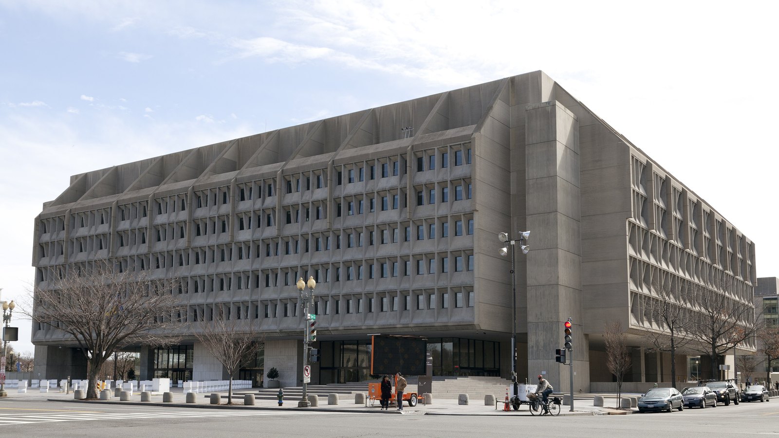 The Hubert H. Humphrey Building in Washington, a long concrete government office block on a corner under a pale winter sky, with parked cars and a cyclist on the street in front of it.