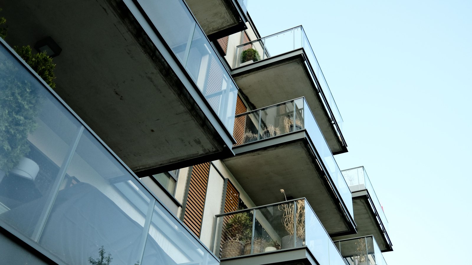 Concrete balcony slabs and glass railings on the side of a modern multi-story residential building, seen from below against a clear sky, with weathering visible on the underside of the balconies.
