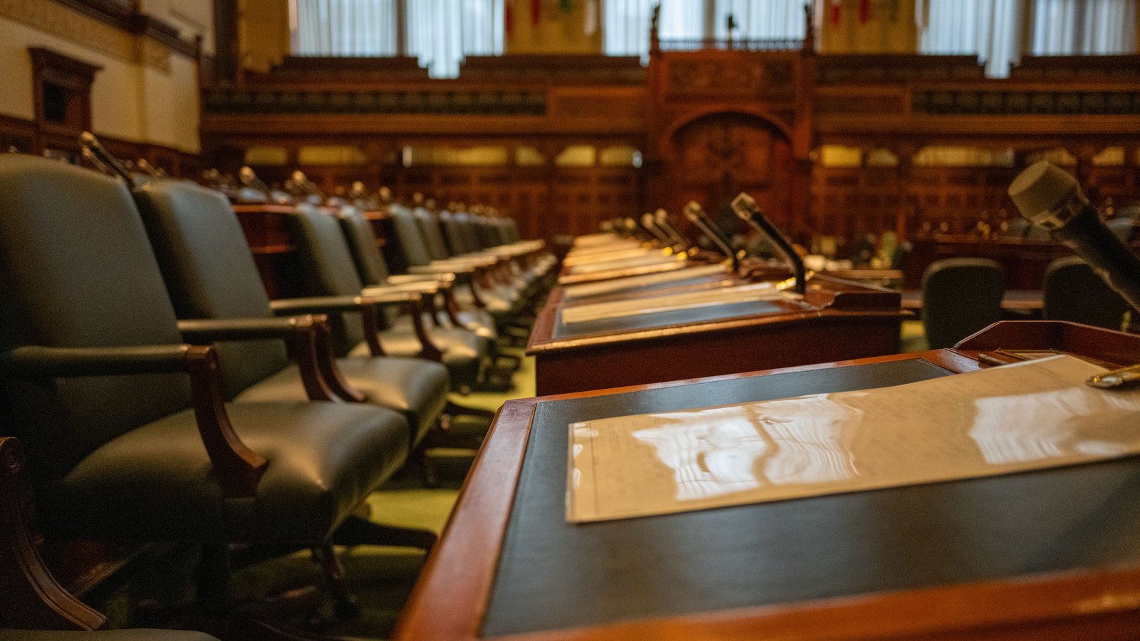 Rows of empty desks in a legislative chamber, each with a microphone and a sheet of paper, green leather chairs pushed in, the far end of the room out of focus.