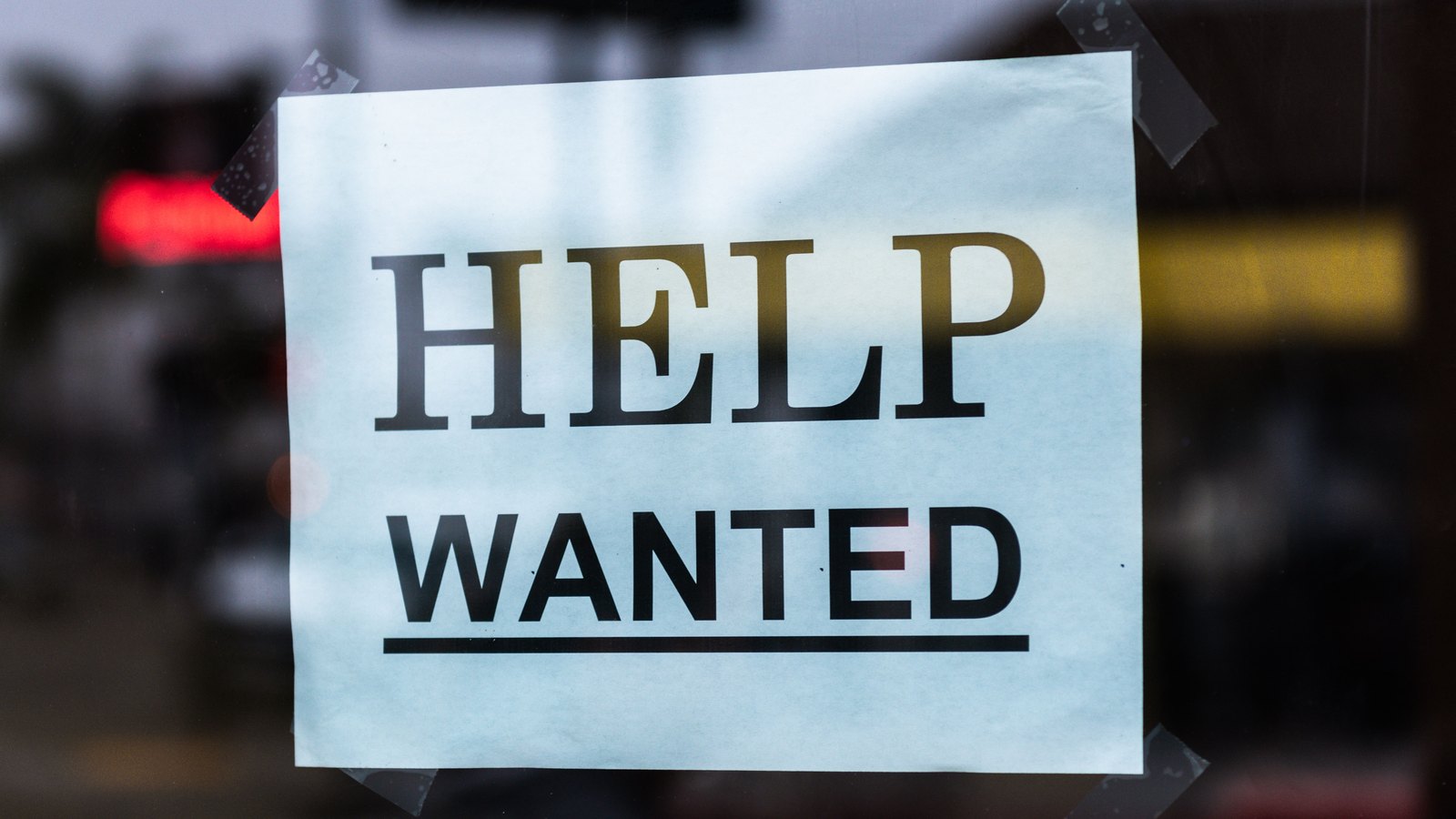A paper HELP WANTED sign taped to the inside of a shop window, with a blurred street reflected in the glass.
