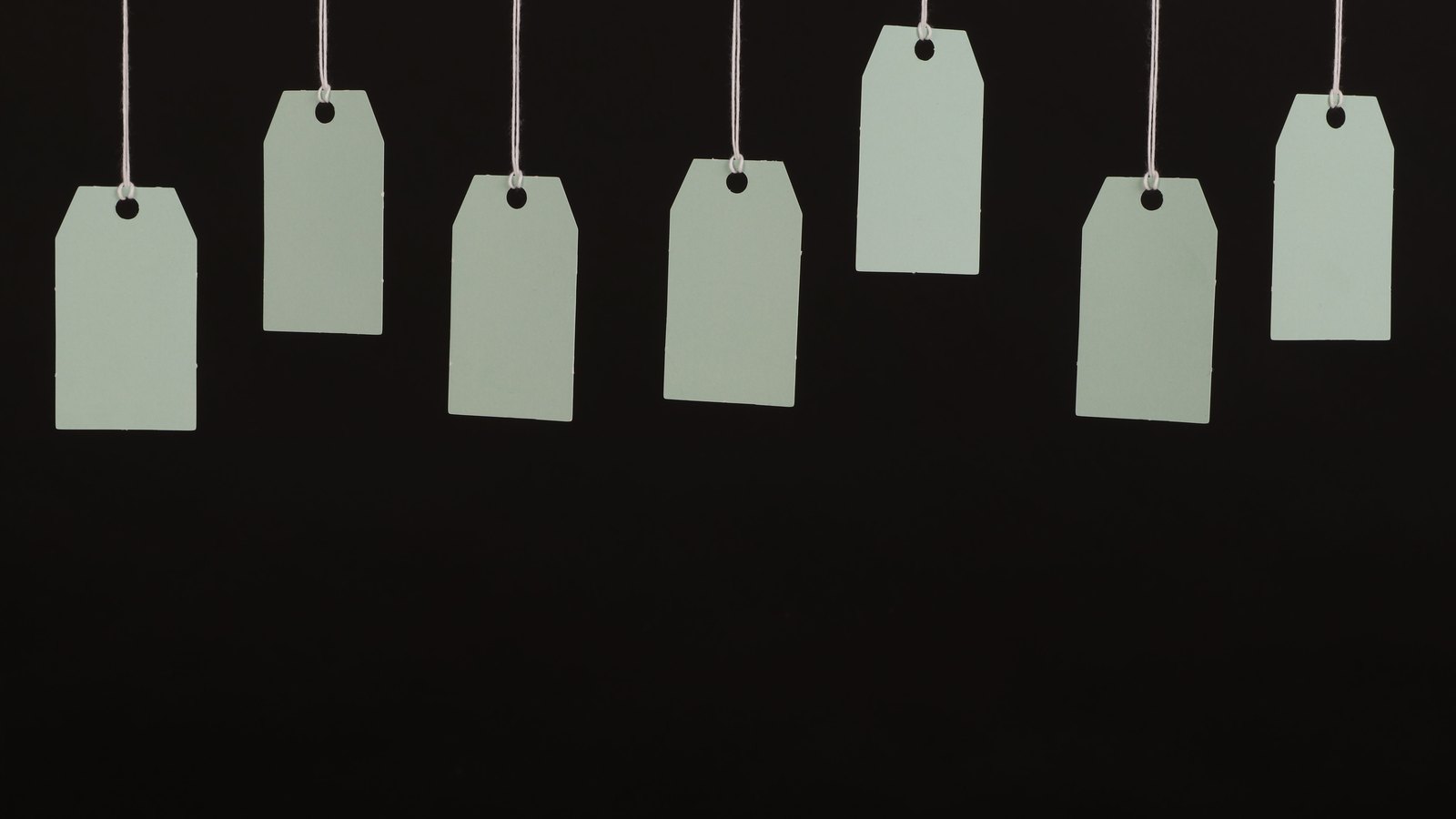 Seven blank paper tags hanging on strings against a black background.