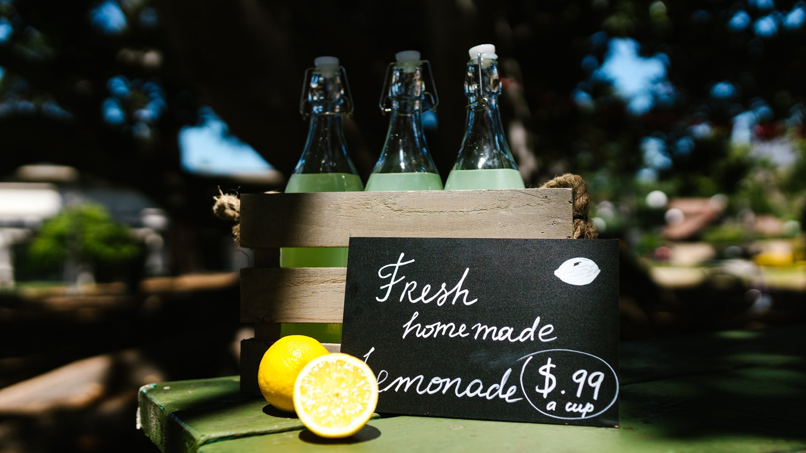 A handwritten chalkboard sign propped against a wooden crate of bottled lemonade, showing a price of ninety nine cents a cup.