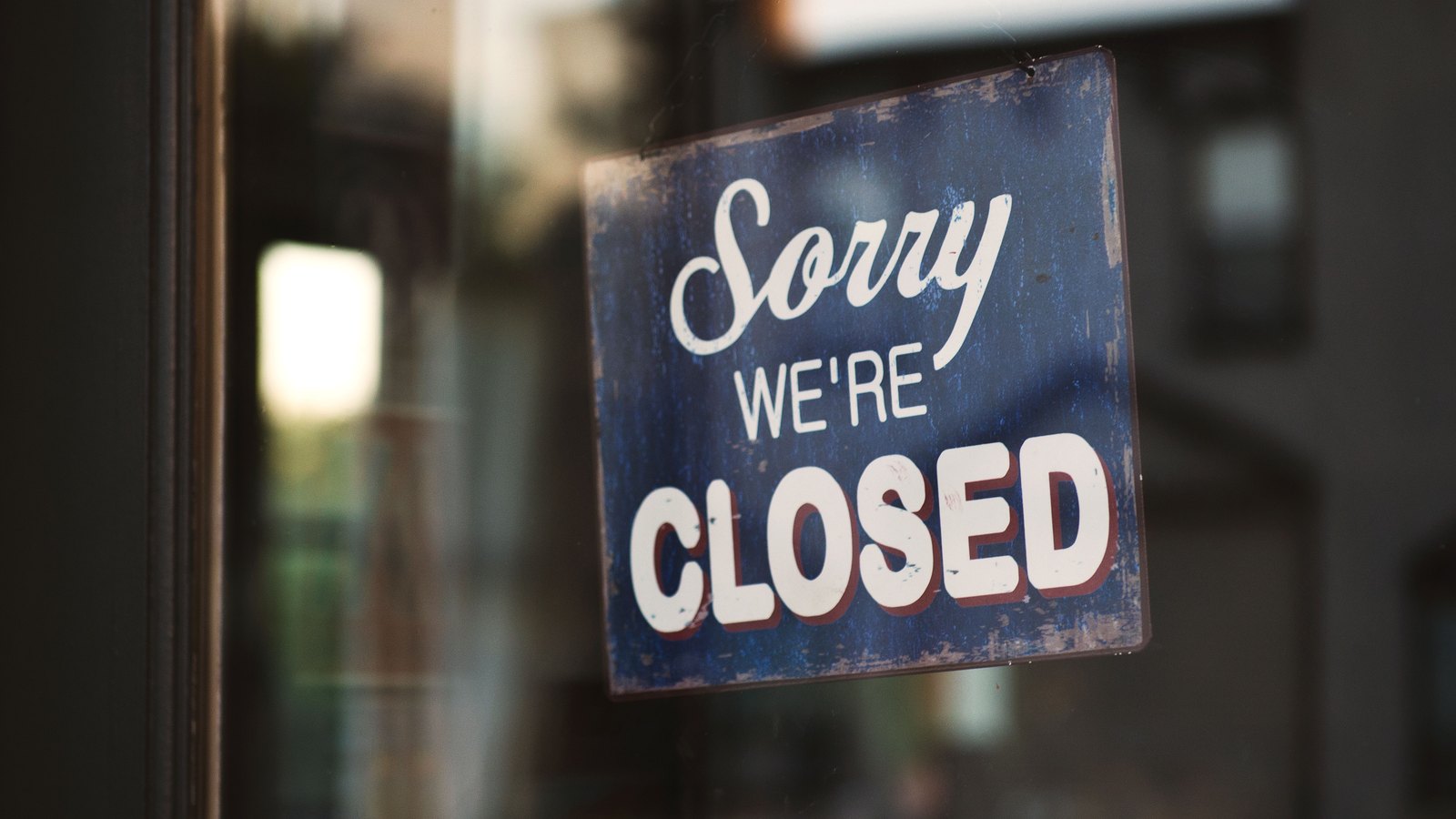 A weathered blue and white metal sign reading Sorry We're Closed hangs inside the glass door of a shop, with the street and buildings opposite reflected in the glass.