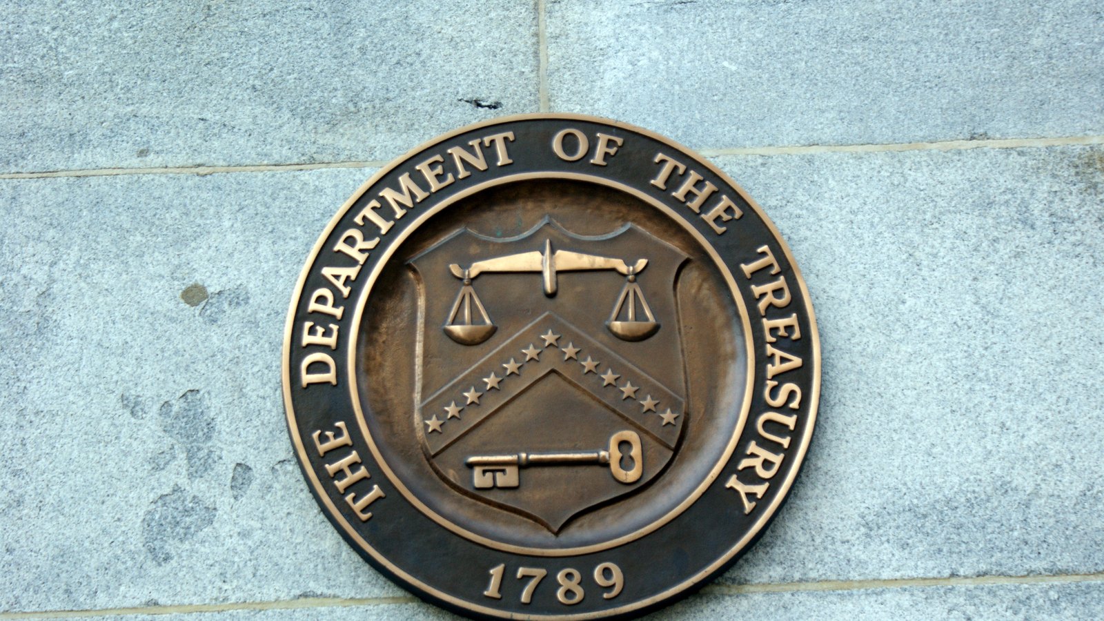 The bronze seal of the Department of the Treasury, showing scales, a key and thirteen stars above the year 1789, mounted on the granite wall of the Treasury building in Washington.