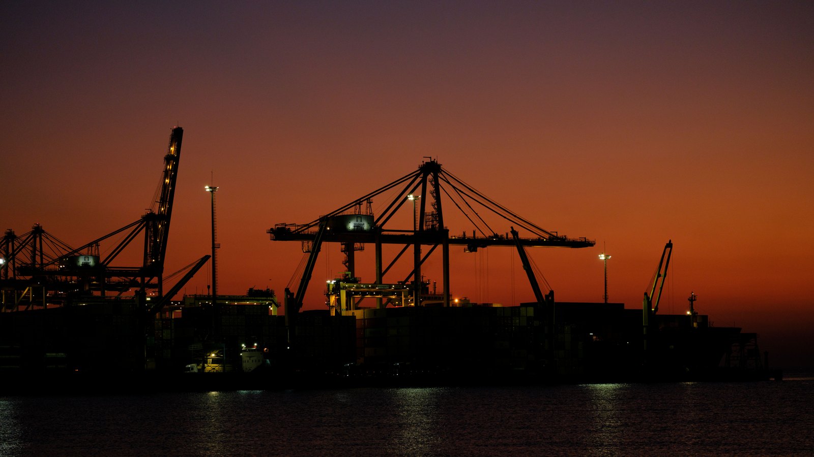 Container gantry cranes and a loaded ship in silhouette against an orange sky at dusk, with dark water in the foreground.