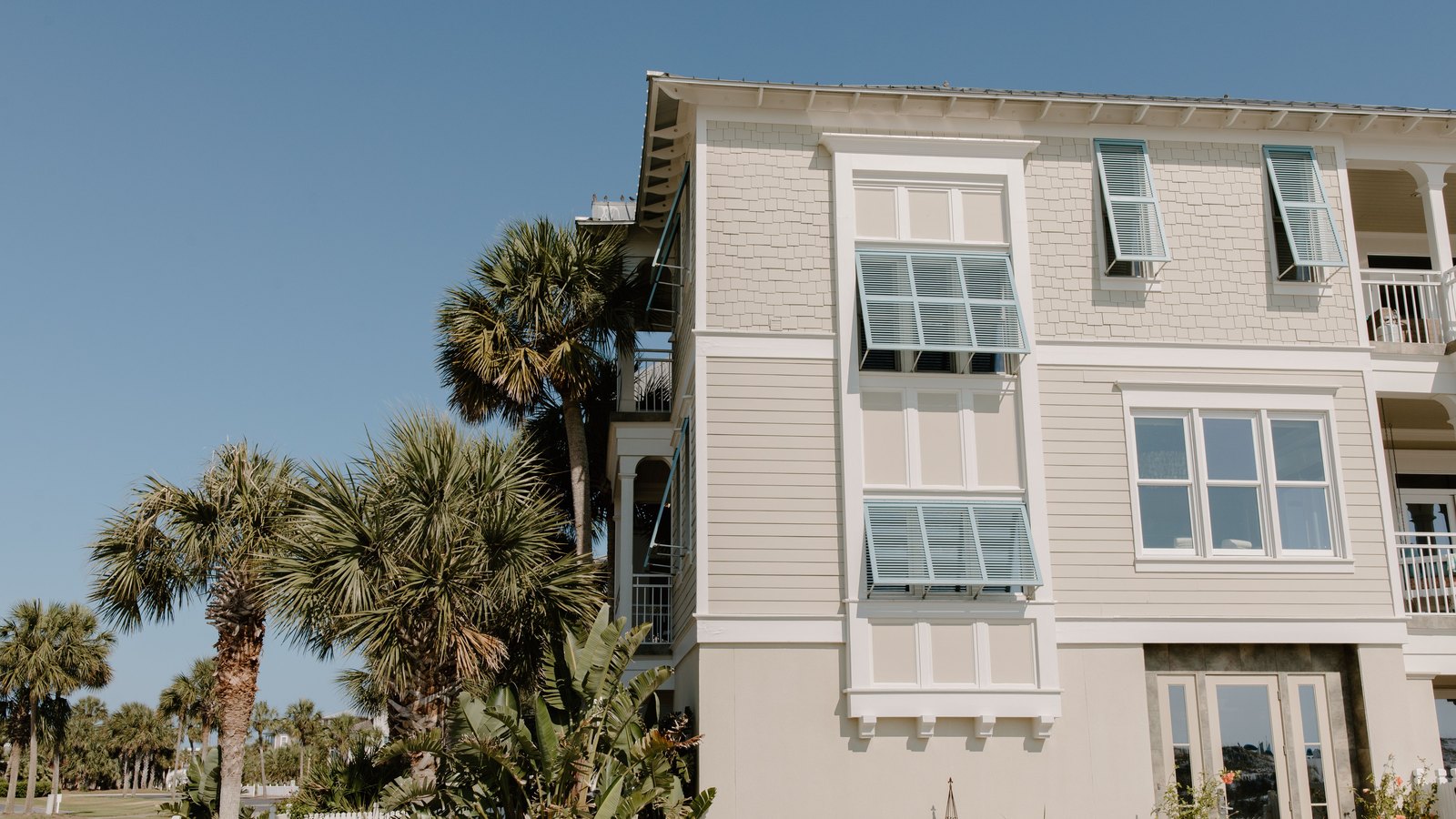 A pale coastal house with palm trees and exterior window awnings
